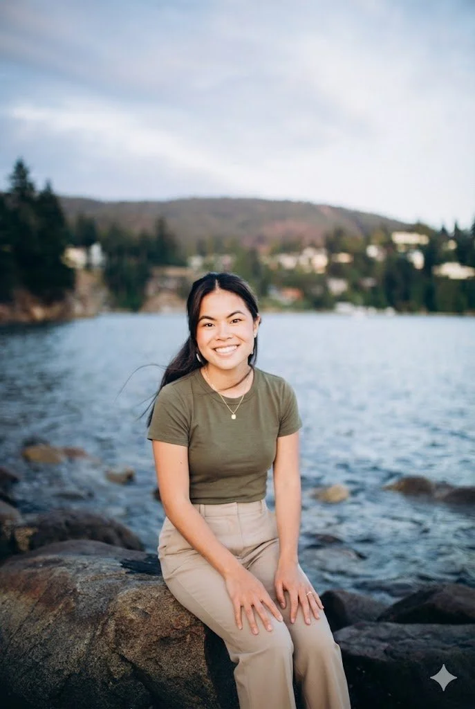 A woman sitting on rocks by a body of water with trees and houses in the background, smiling at the camera.
