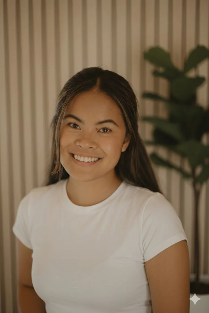 A young woman with long dark hair, smiling, wearing a white t-shirt, standing in front of a background with vertical beige and white stripes and a large green plant.