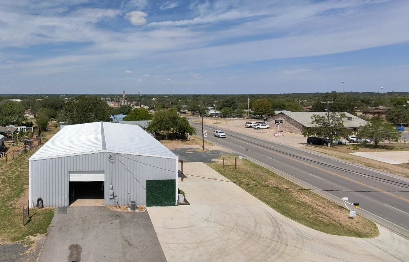 A large metal building with a white roof and gray walls sits on a concrete lot next to a paved driveway in a small town. A bus stop symbol is visible on a sign beside the sidewalk. The street in front has a few vehicles driving by, with trees and several other buildings in the background under a partly cloudy sky.