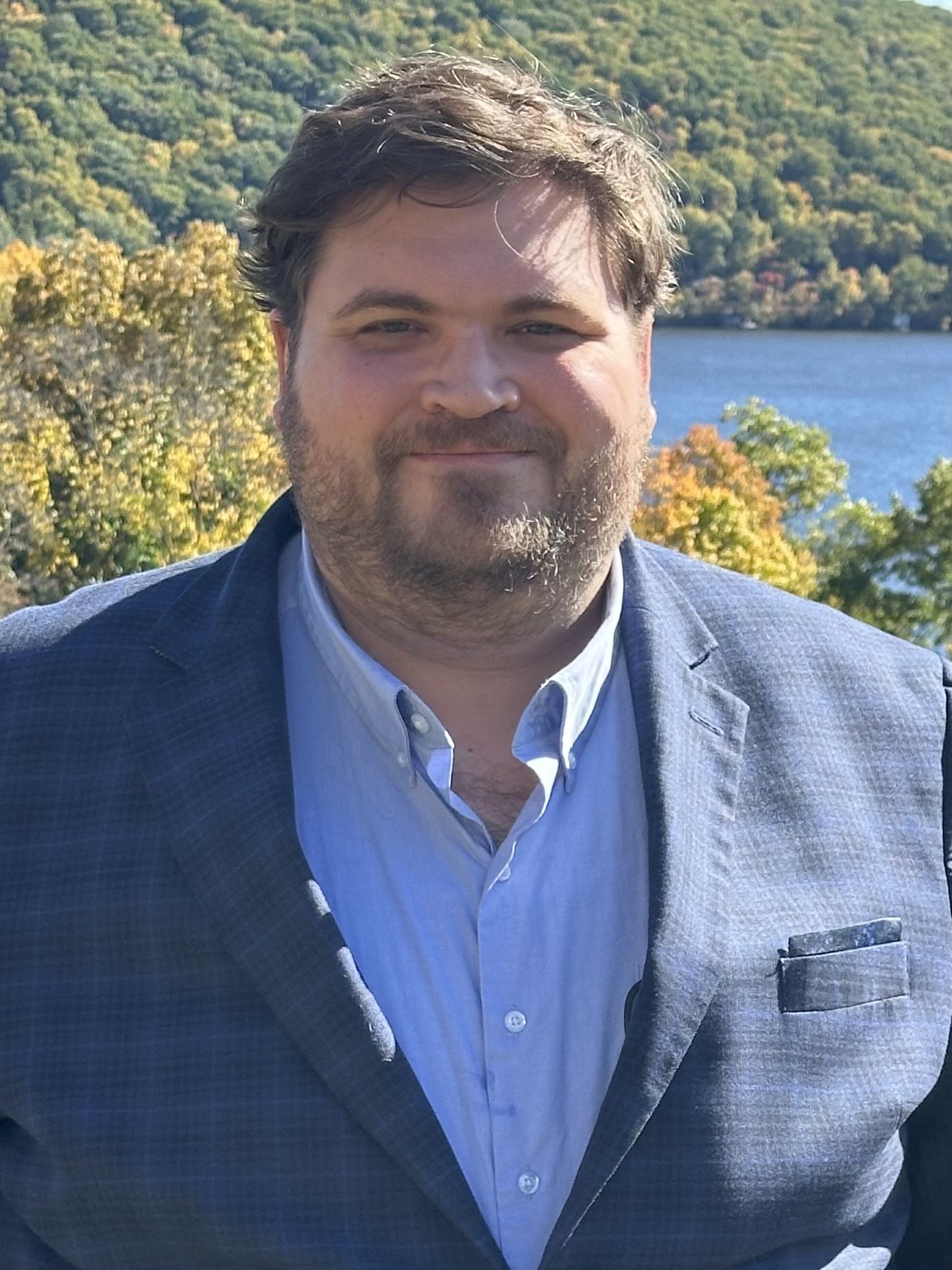 Male presenting figure, in sports coat and blue, facing camera and smiling.