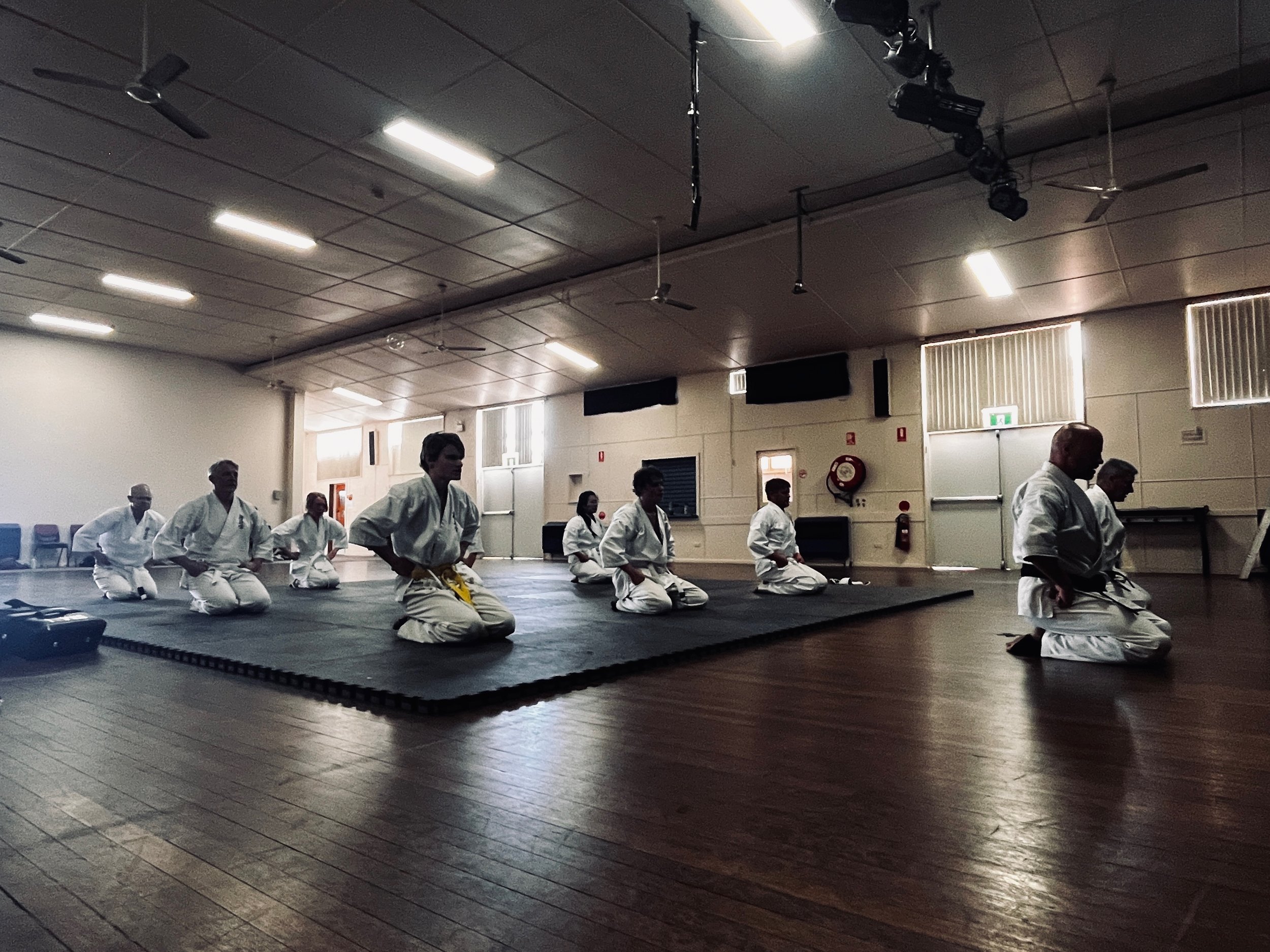 Martial arts class with students kneeling on mats, wearing white uniforms, in a spacious indoor training room with wooden floors and ceiling fans, some standing in the background.