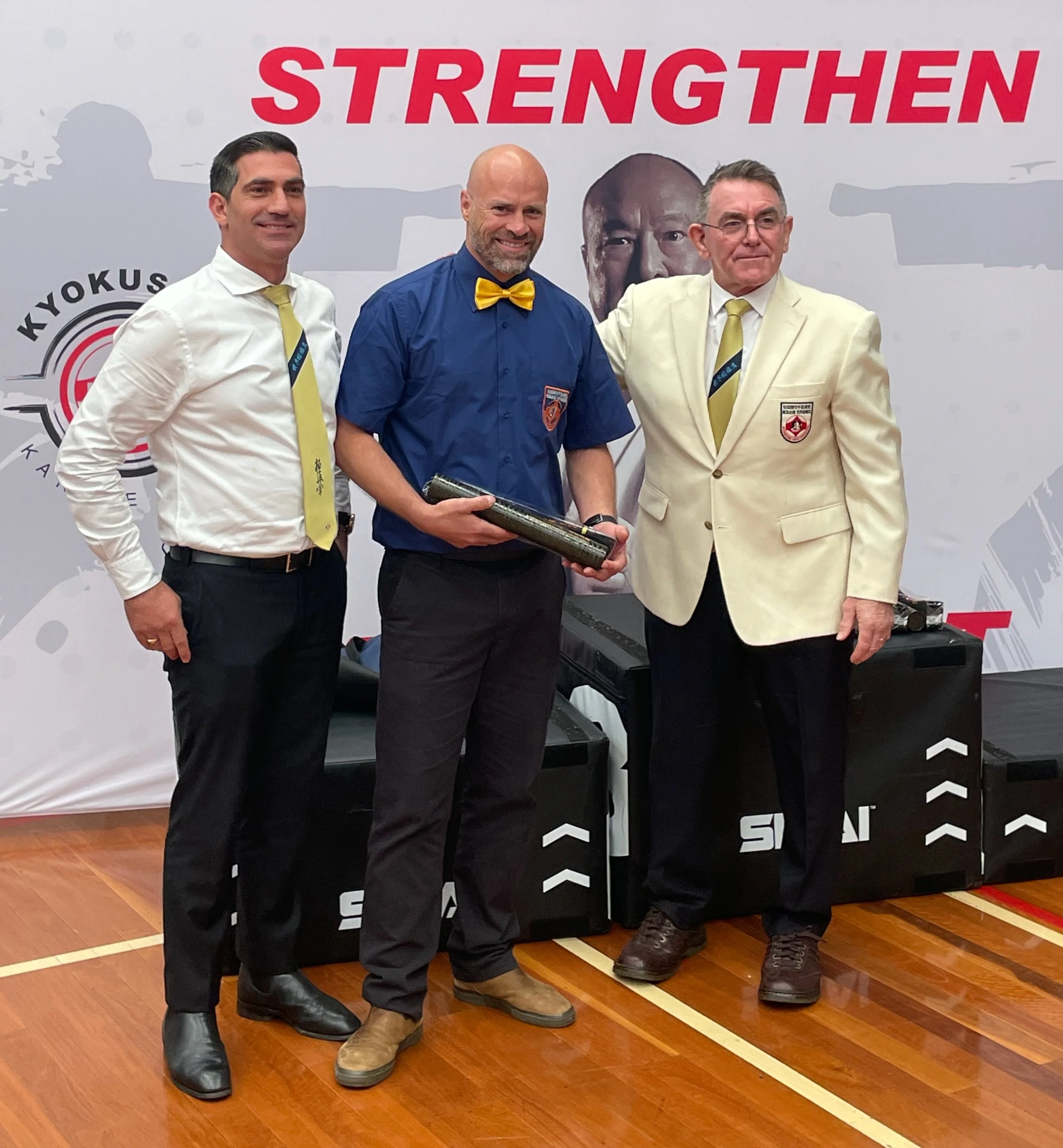Three men standing together on a wooden floor, with a backdrop that has the word 'STRENGTHEN' in large red letters and the Kyokushin Karate logo. The man in the middle is wearing a blue shirt with a yellow bow tie, holding a certificate or award. The men on the left and right are dressed in white suits with yellow ties, and the man on the right has a patch on his blazer. They are posing for a photo.