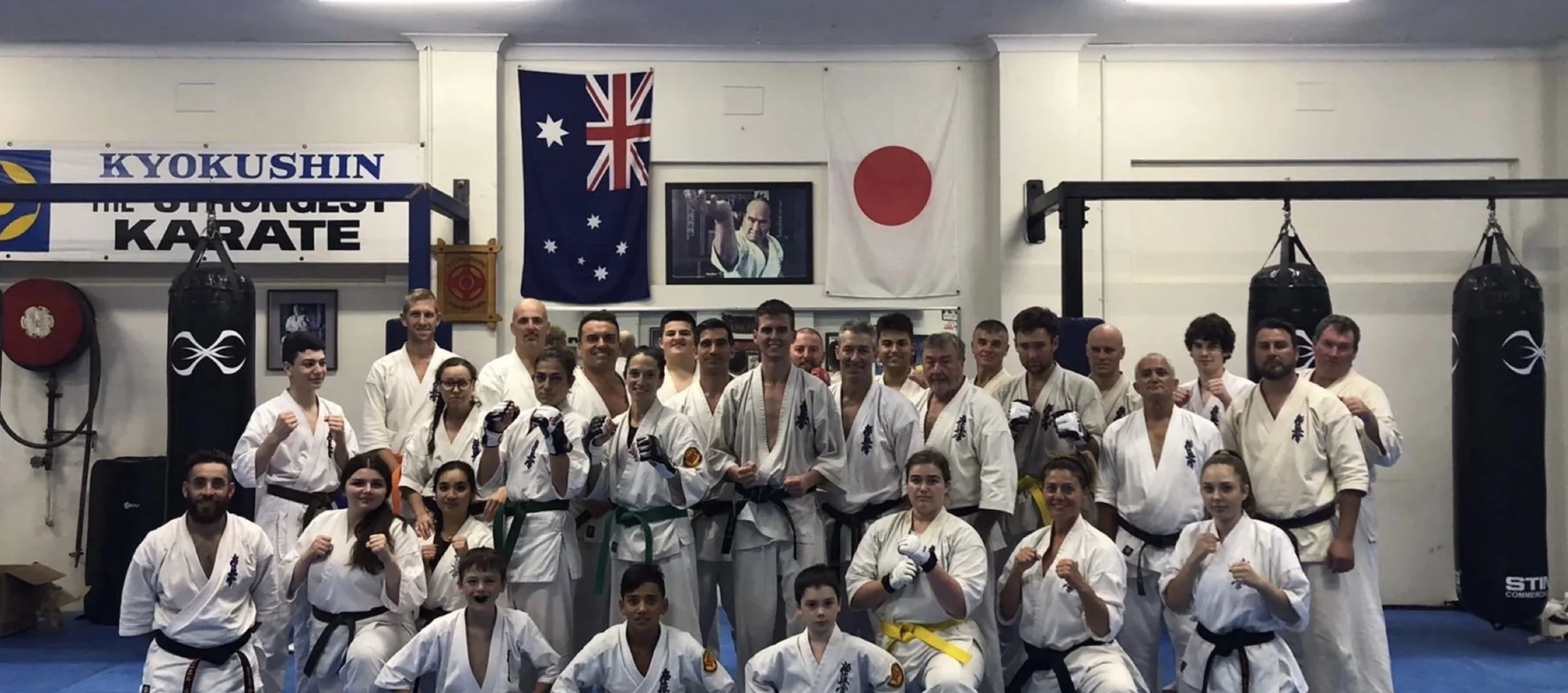 Group of martial artists in karate uniforms posing for a photo in a dojo with flags and punching bags in the background.