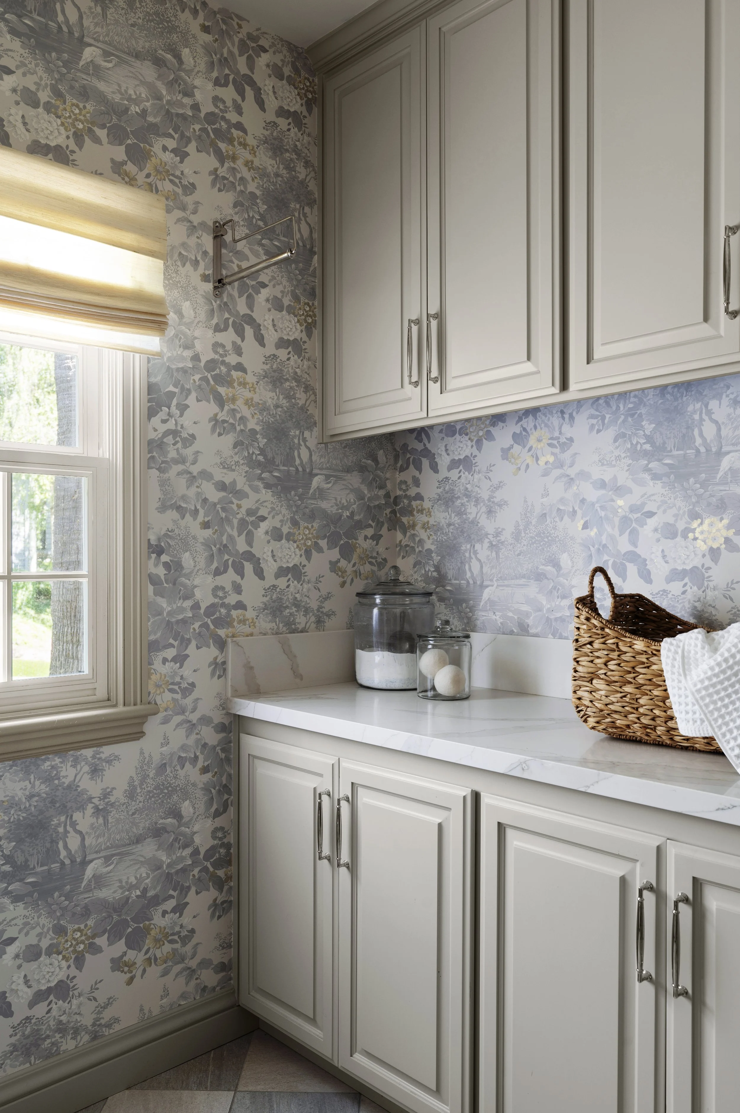 A kitchen countertop with decorative jars, a wicker basket, cabinets, patterned wallpaper, and a window with a beige shade.