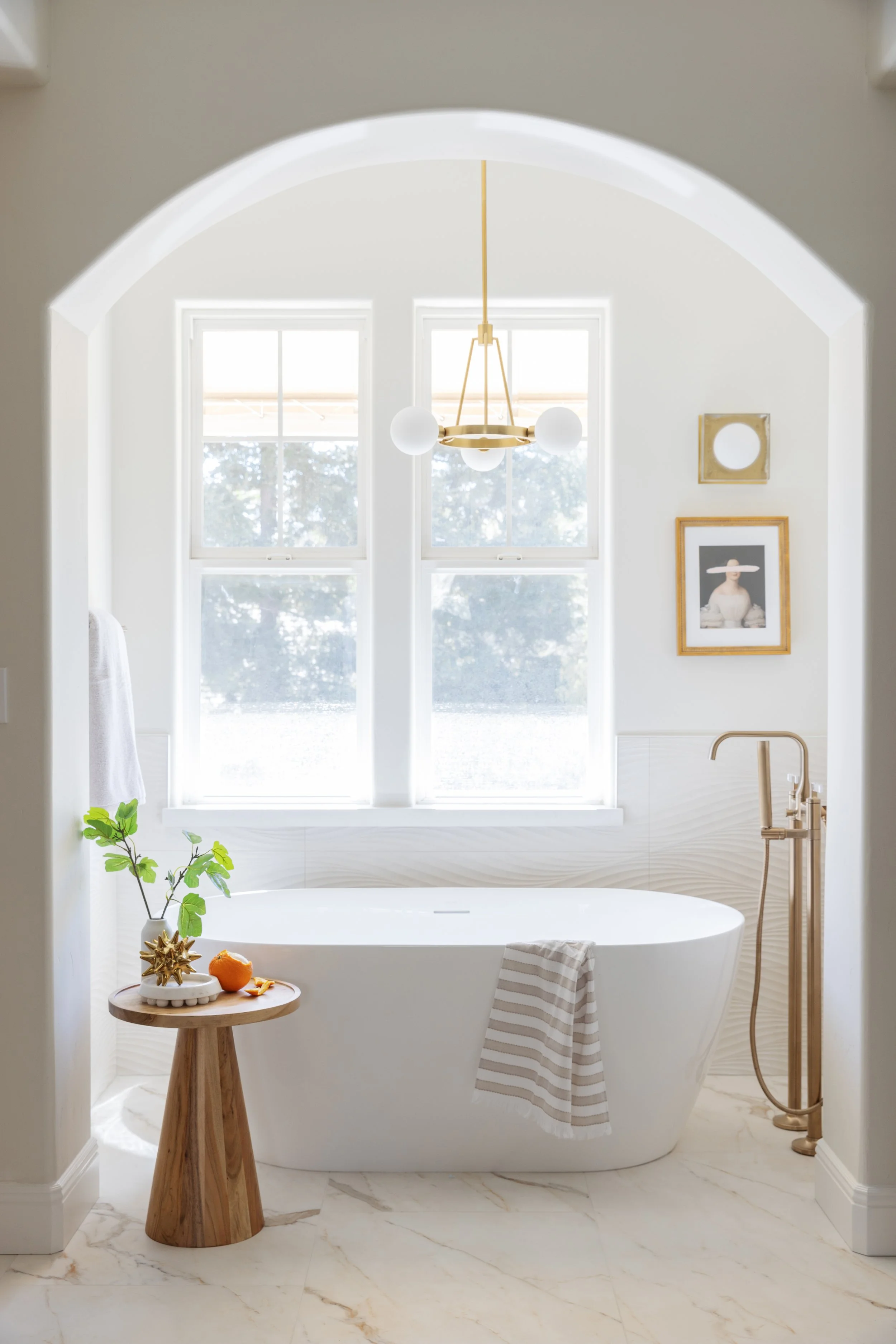 Bright bathroom with a freestanding bathtub near two large windows, a small wooden side table with decorative items, and a towel hanging on the side of the bathtub. The room features light-colored marble flooring, a modern chandelier, and wall art.
