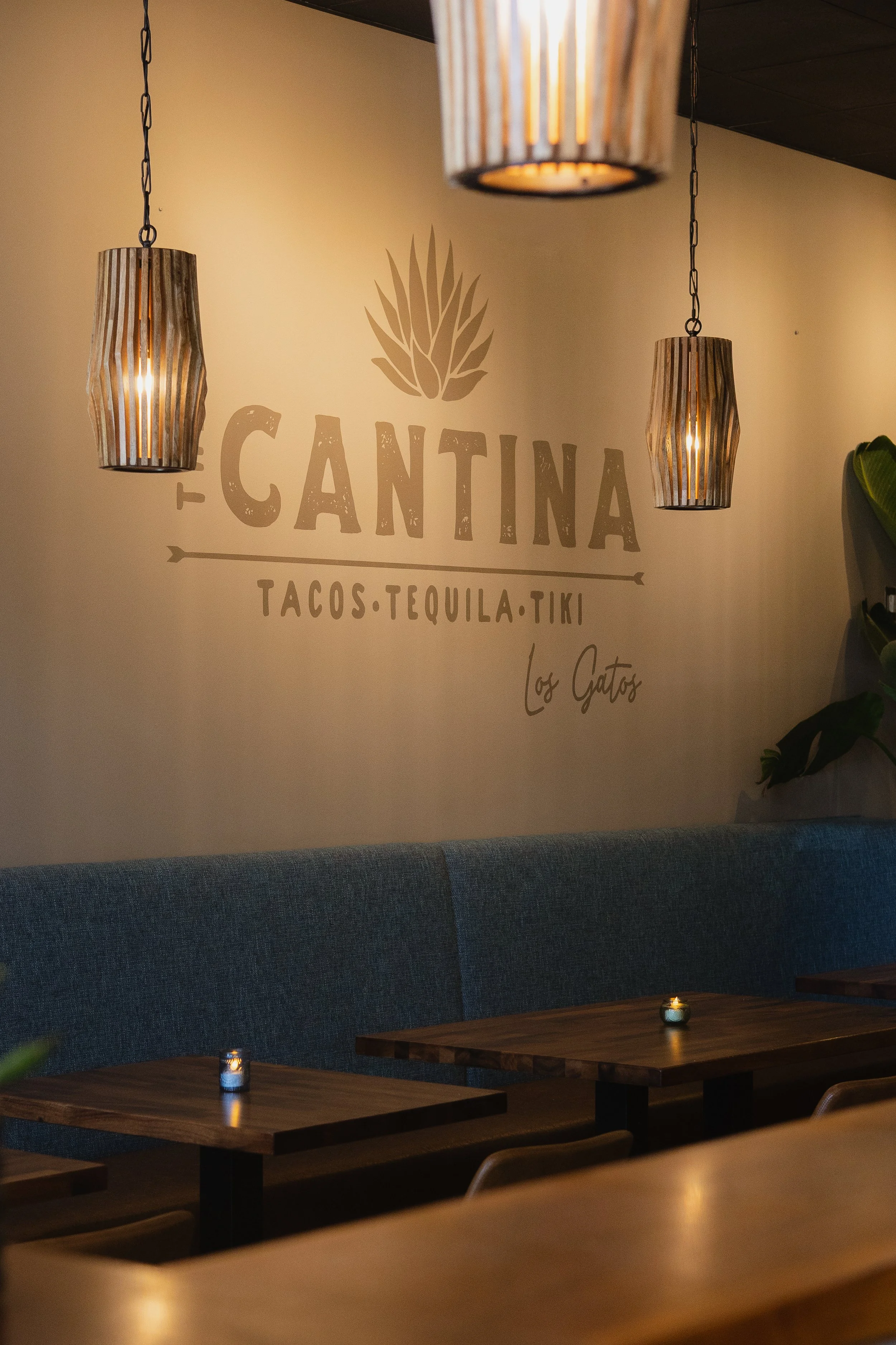 Interior of a restaurant with hanging wooden pendant lights, a beige wall with the words 'Cantina Tacos Tequila Tiki' and a decorative leaf graphic, dark blue upholstered seating, and wooden tables with small lit candles.