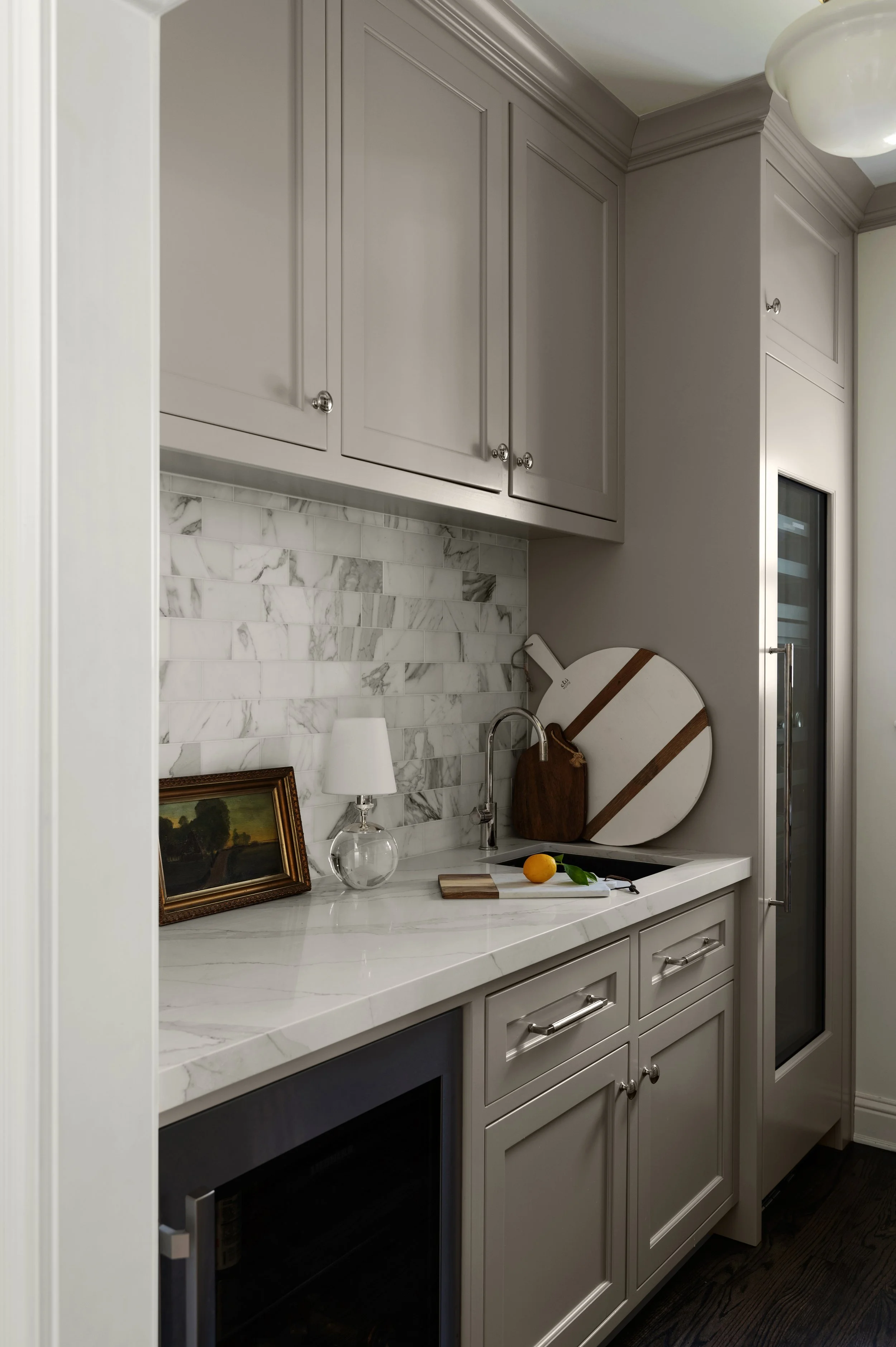 Kitchen with gray cabinets, white marble backsplash, marble countertop, small lamp, framed painting, glass vase, cutting board with lemon, and a cutting knife, microwave oven, and a door with a wine cooler built-in.