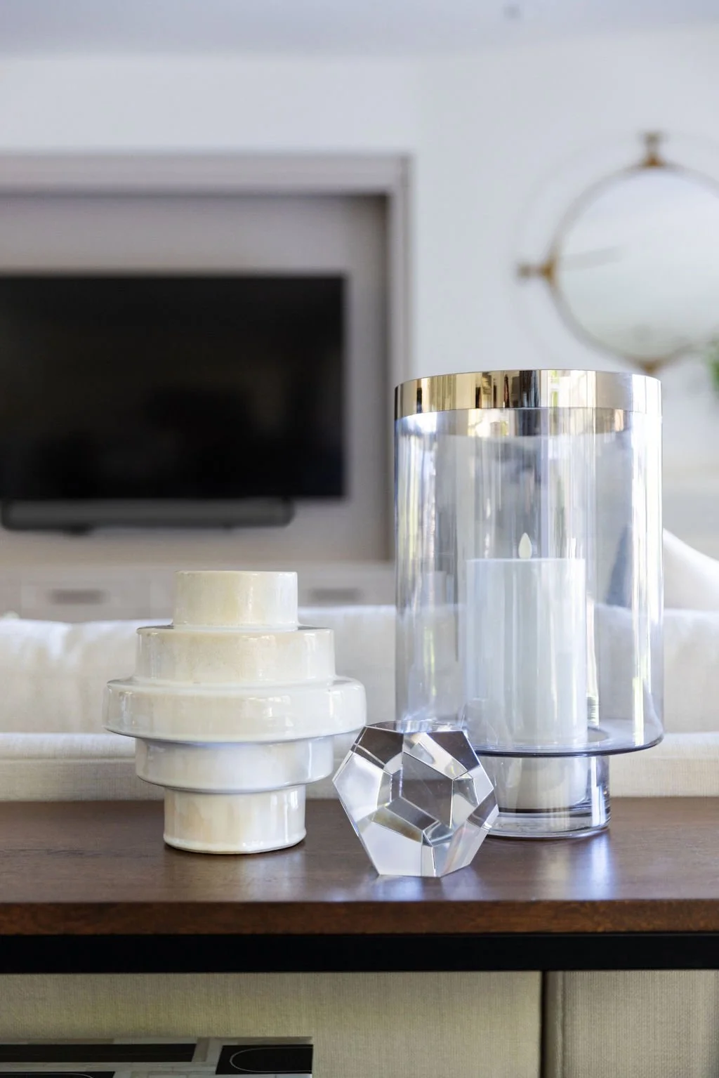 Decorative objects on a wooden table, including a white tiered ceramic piece, a large glass hurricane with a candle inside, and a multifaceted glass sculpture, with a blurred television and clock in the background.
