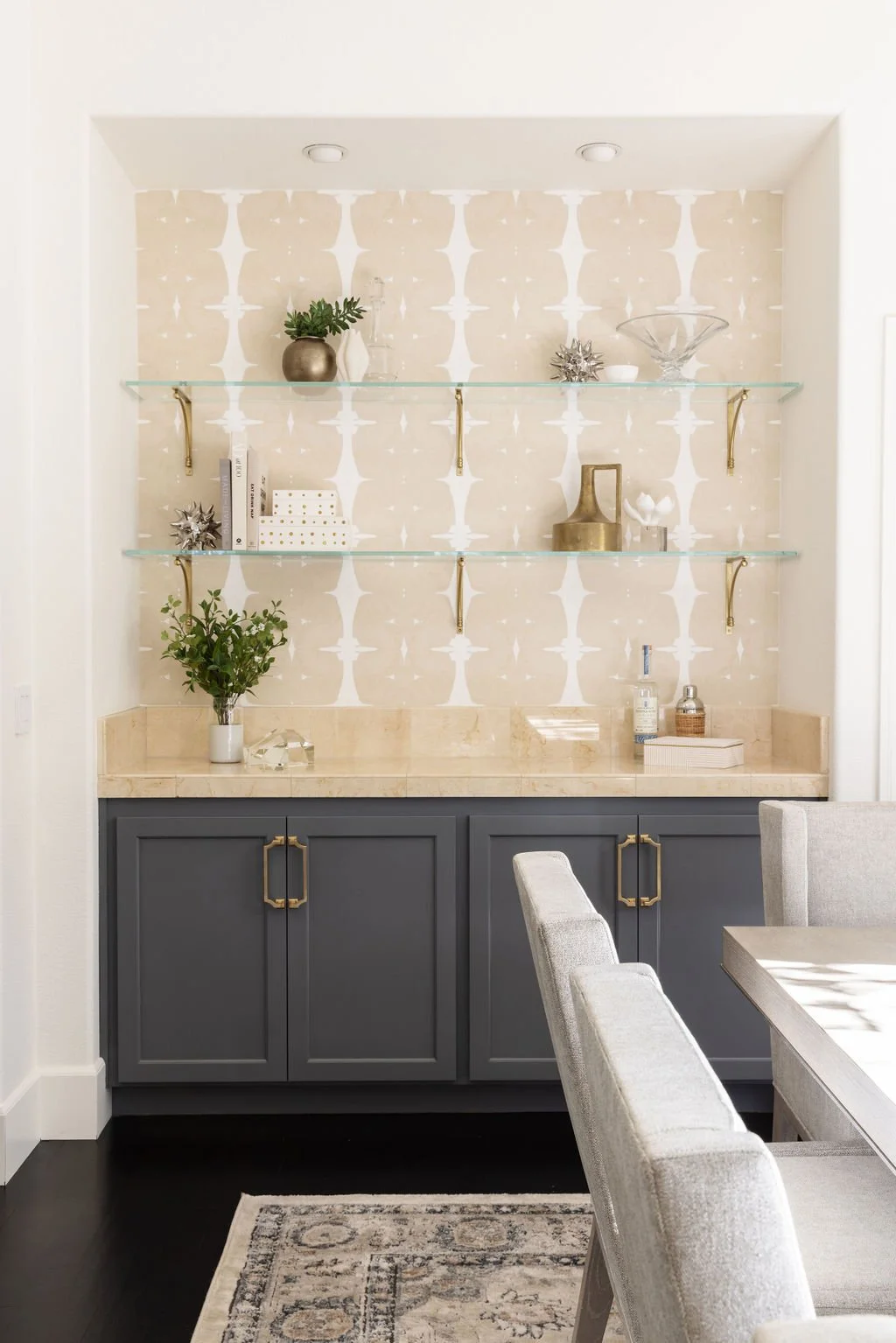 Interior of a modern dining room featuring a built-in display niche with glass shelves, decorated with vases, ornaments, and books. The bottom cabinetry is dark gray with brass handles, and the countertop is beige marble. A dining table with upholste