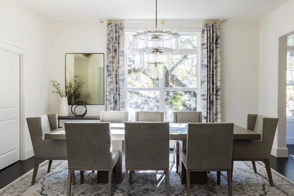 Modern dining room with white walls, large window with patterned curtains, chandelier, rectangular table, tan upholstered chairs, decorative items, and a mirror.