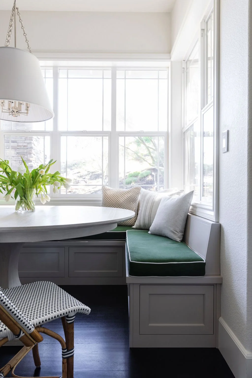 A cozy dining nook with a round white table, green cushion bench, and a window seat with pillows. A vase of white flowers on the table, and a large white dome pendant light hanging from the ceiling.