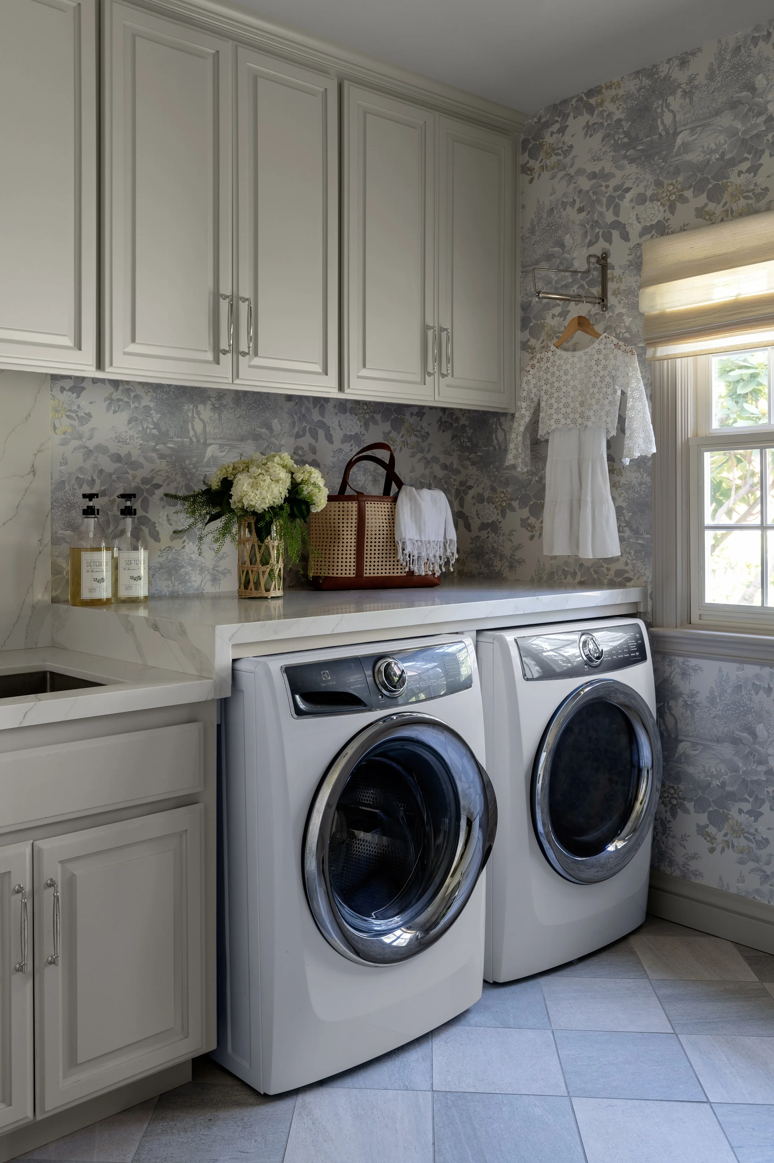 A laundry room with a white countertop, beige cabinets, floral wallpaper, a window with beige roman shades, and a pair of front-loading washing machines. Decor includes a vase of white flowers, a woven basket, a white towel, and a white dress hanging on a wall hook.