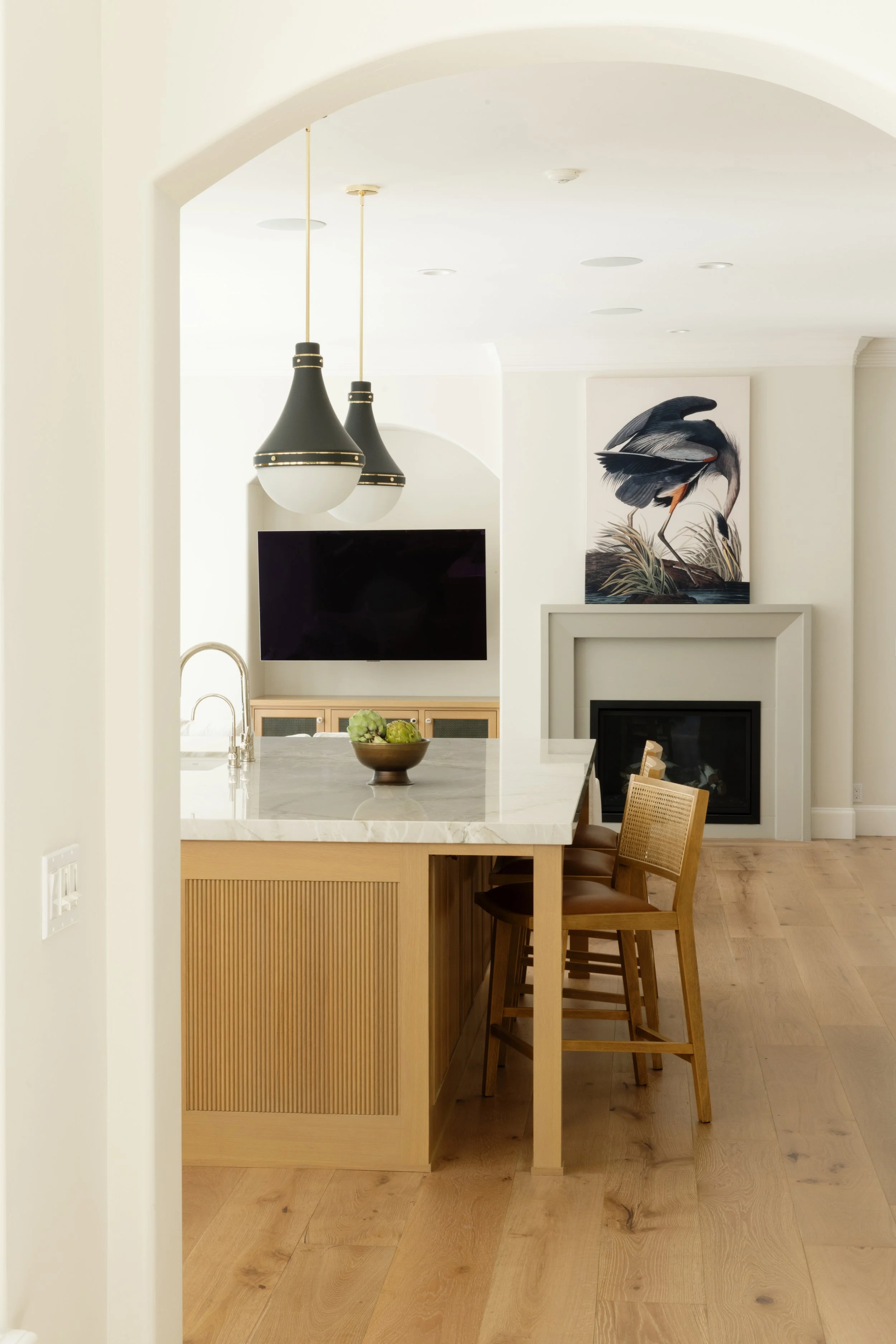 A kitchen with a marble island, bare wooden chairs, a bowl of artichokes, a wall-mounted TV, two black pendant lights, a fireplace, and a large heron artwork in the background.