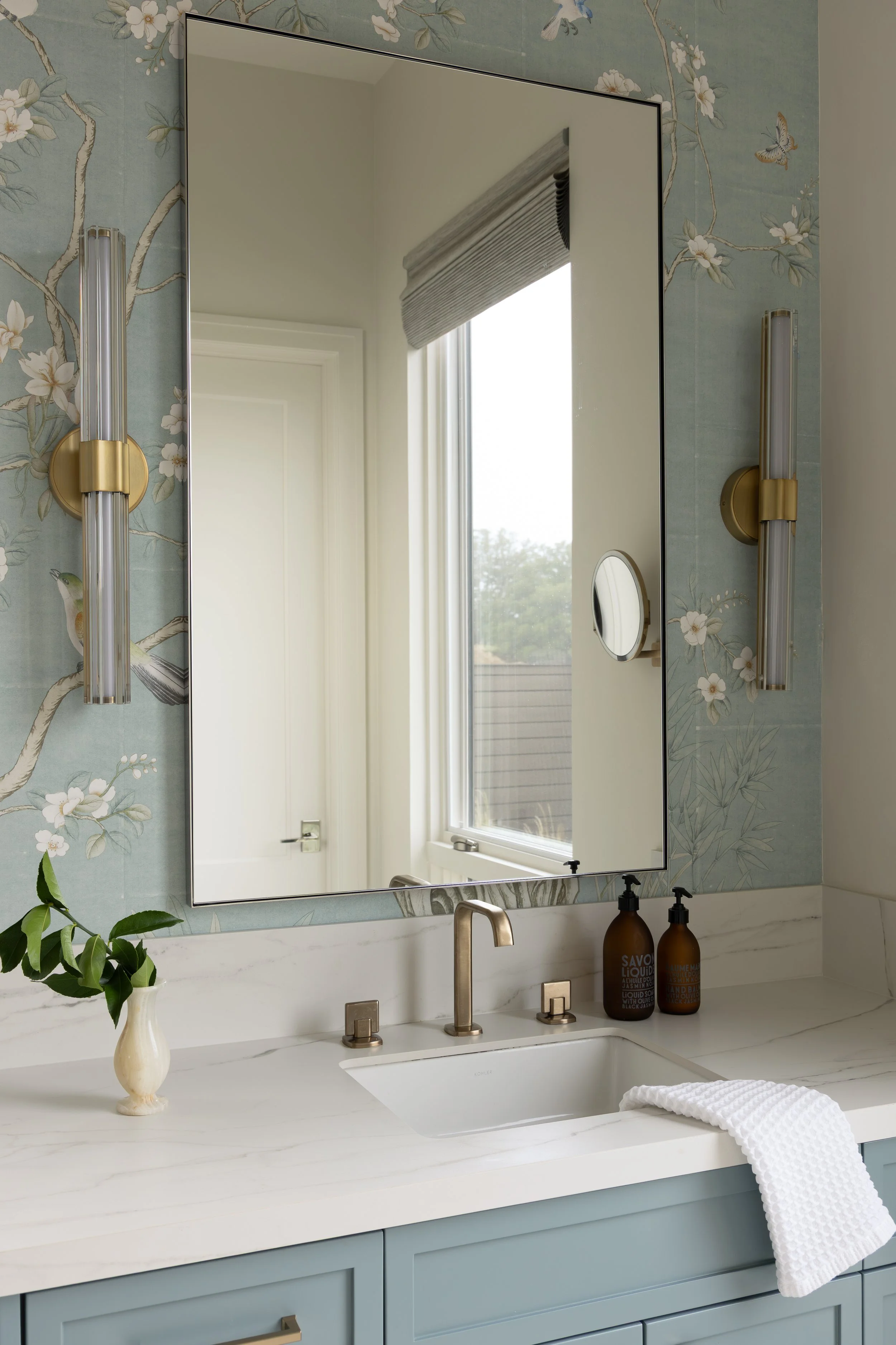 A bathroom vanity with a white marble countertop, a white undermount sink, and a gold faucet. Two brown bottles of liquid soap or lotion are on the counter. A white textured towel is draped over the edge of the counter. To the left, a white vase with green leaves. The wall has floral wallpaper with white flowers and branches, and a bird. A large mirror with slim black frame is above the vanity, reflecting a window with blinds and a door.