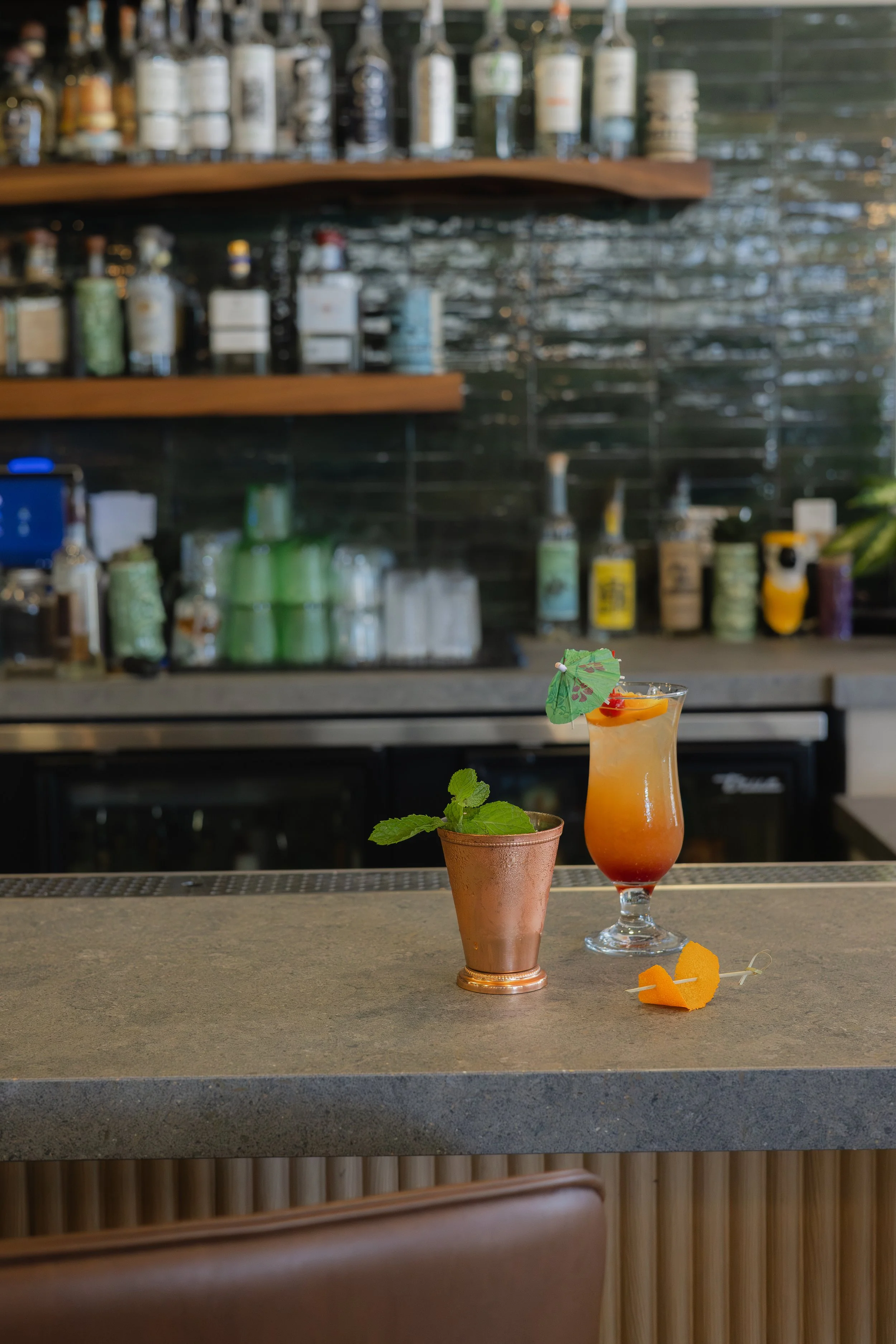 A colorful tropical cocktail with a small umbrella and a cherry, garnished with a mint sprig, placed on a bar counter in front of a bar with shelves of liquor bottles.