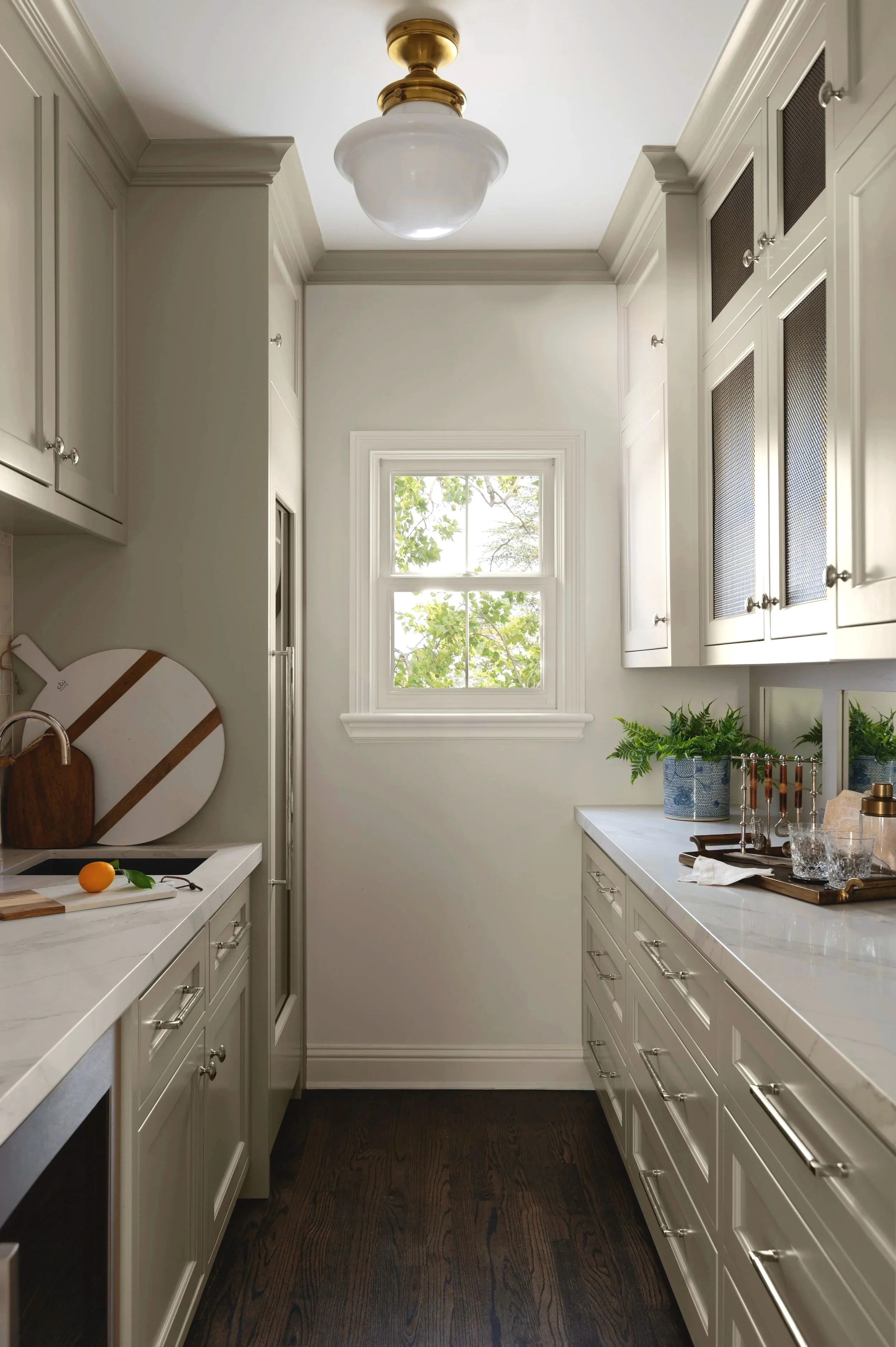A narrow kitchen with white cabinets, a window with green foliage outside, a white countertop, and a dark hardwood floor. The ceiling has a vintage-style light fixture with bronze and glass elements.
