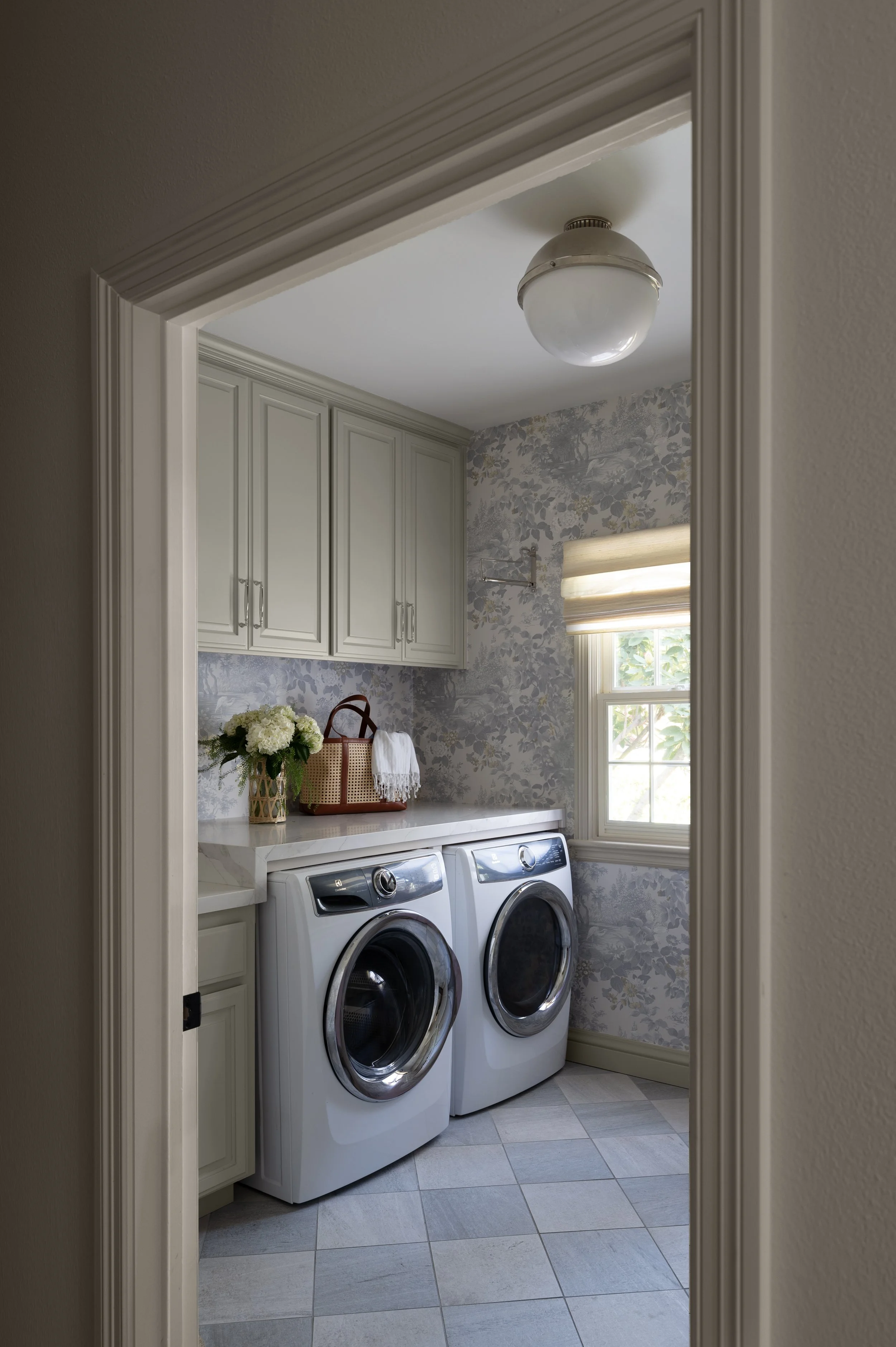A laundry room with a top window, floral wallpaper, white cabinets, a floral vase with white flowers, and a woven basket. There are two front-loading washers and a bathroom fixture visible.