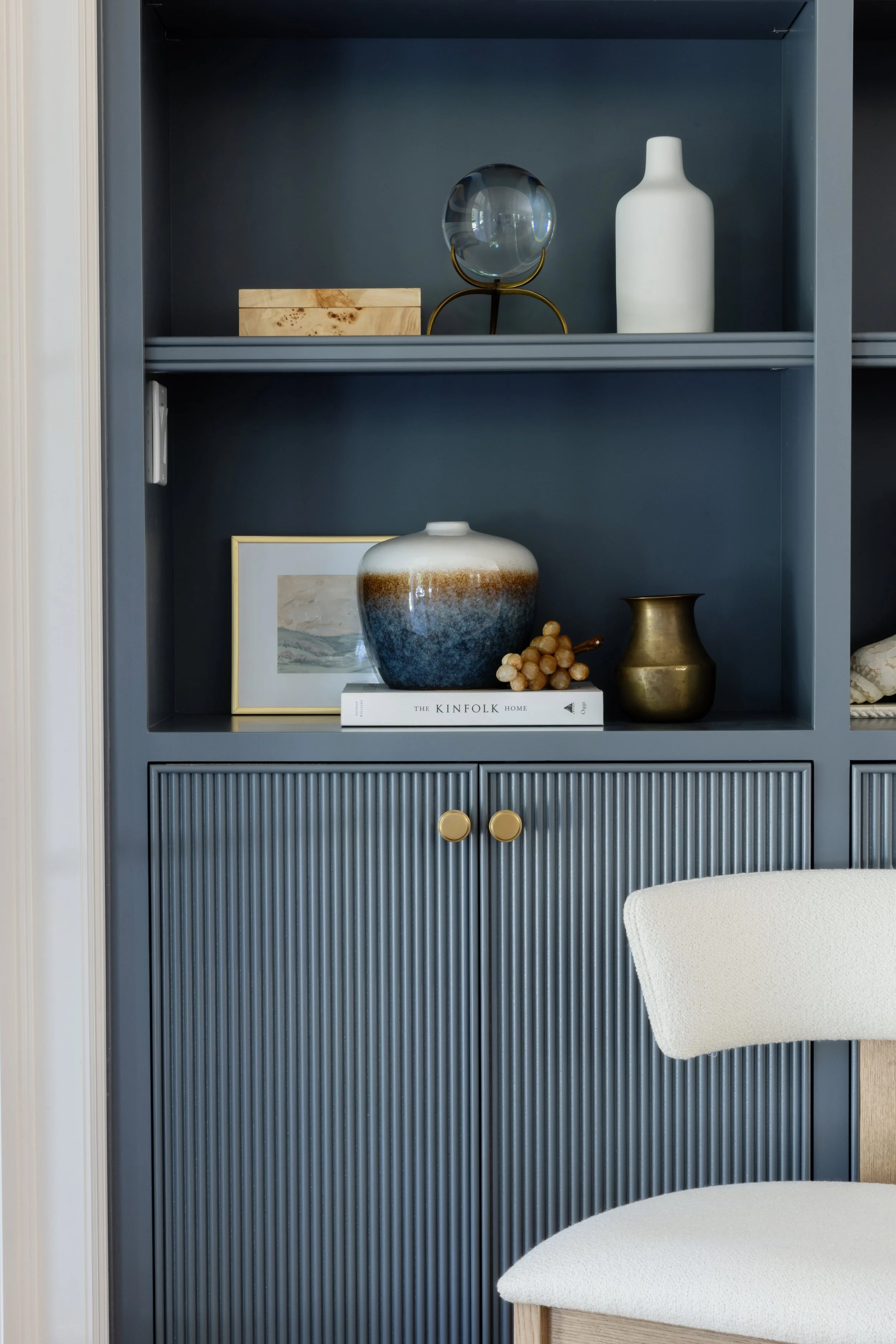 Blue shelving unit with decorative items including a clear glass orb on a brass stand, a white matte vase, and a wooden box. Below, a framed artwork, a large ceramic vase with blue, beige, and white glaze, a small wooden bead arrangement, and a brass