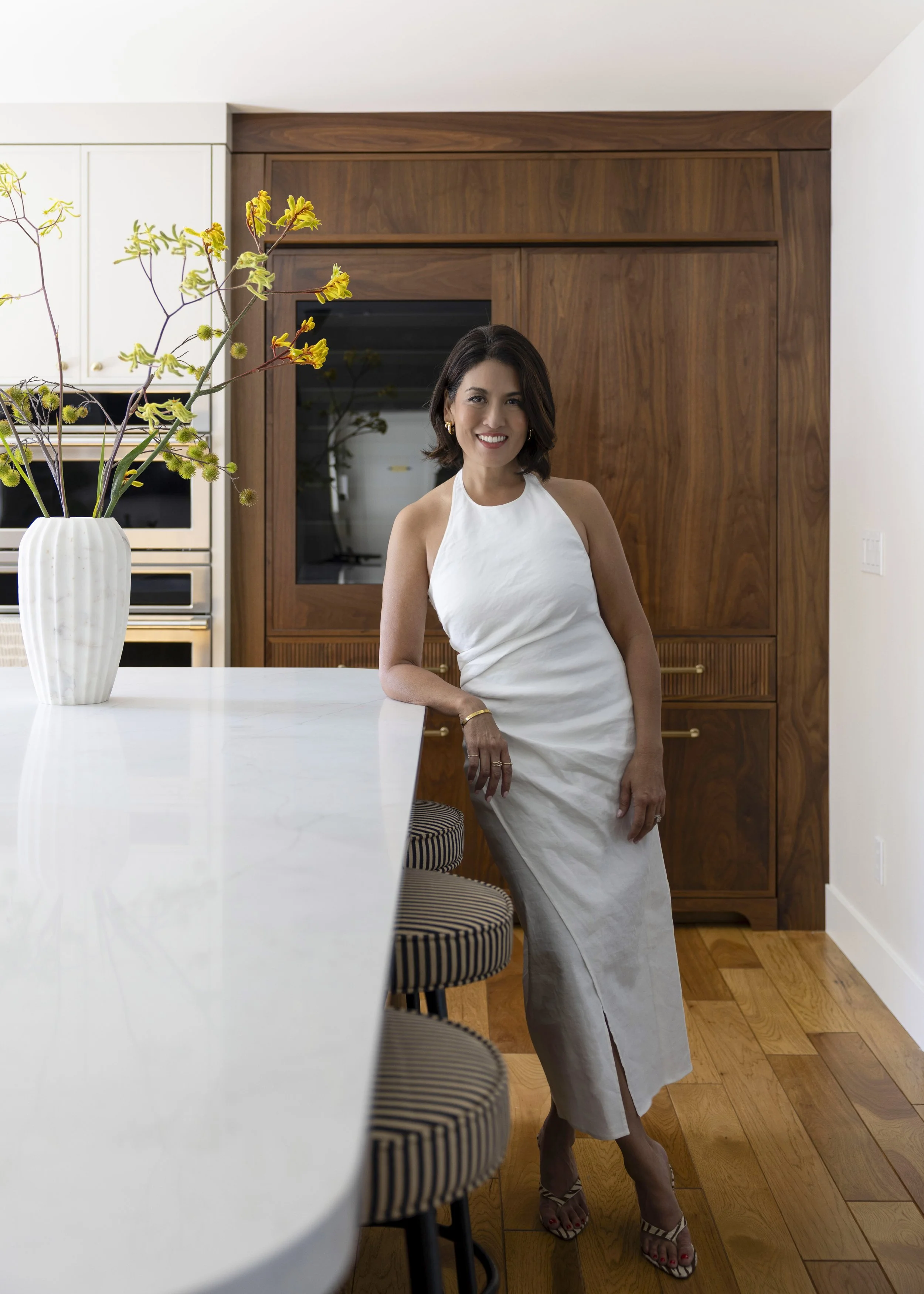 A woman with dark hair, wearing a white sleeveless dress and high heels, standing in a modern kitchen with wood cabinets and a white countertop, smiling at the camera.