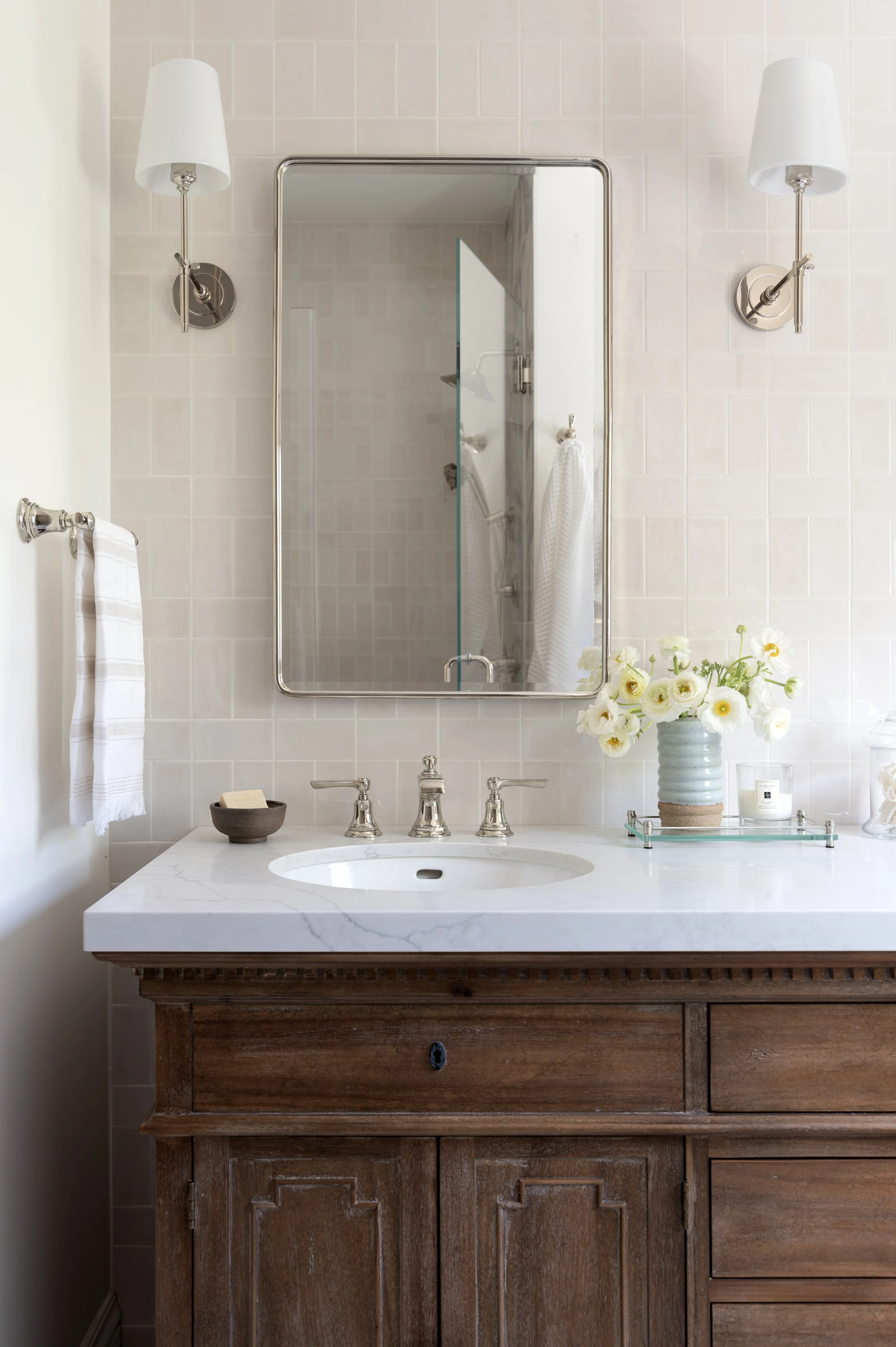 Bathroom vanity with a marble countertop, built-in sink, and a wooden cabinet. Above the vanity is a large mirror flanked by two wall-mounted lamps. On the right side of the countertop, there is a vase with white flowers, a small tray, and a candle. 