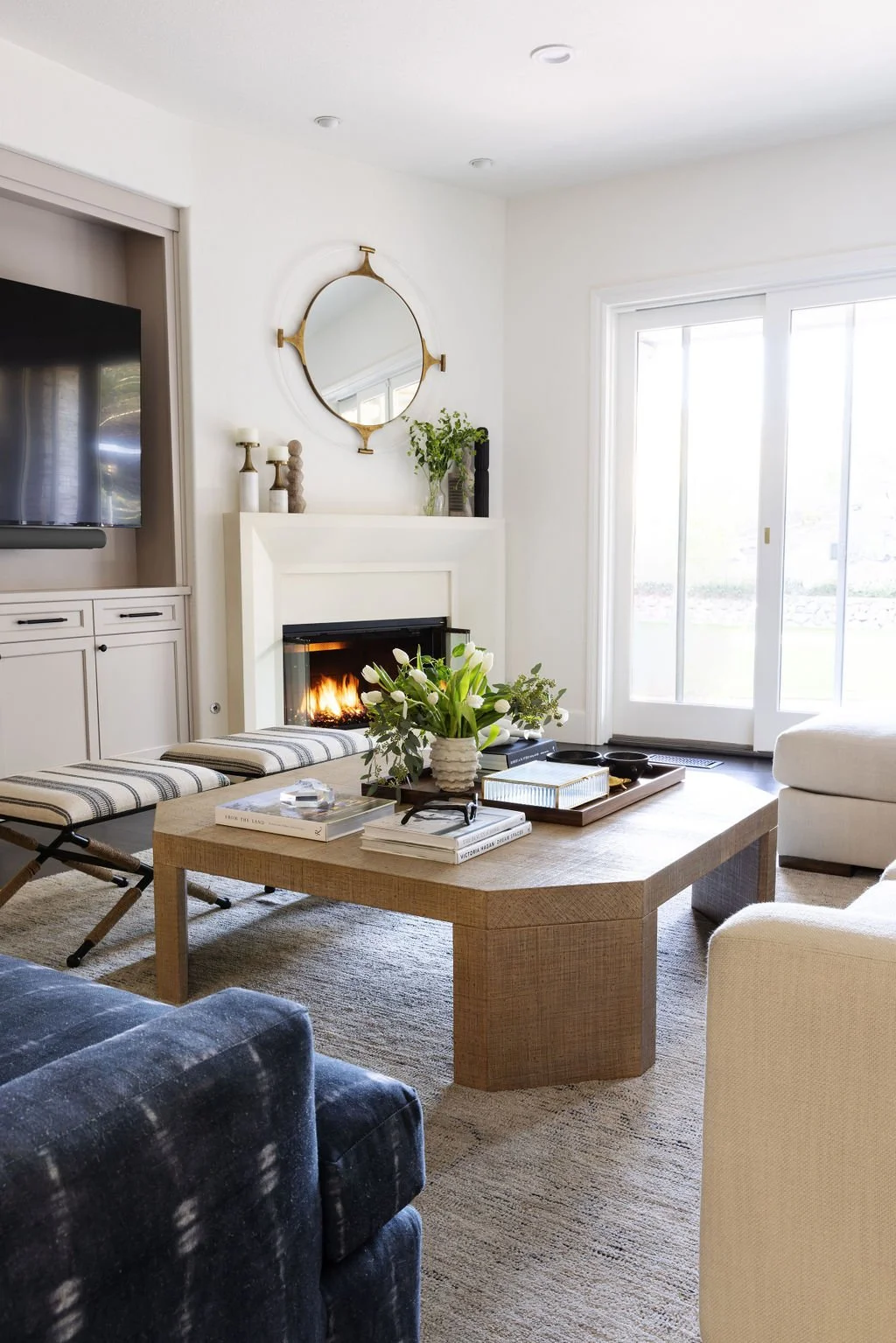 Living room with a wooden coffee table, a fireplace, a sliding glass door, a TV, and a beige armchair. Decor includes flowers, books, and a mirror above the fireplace.
