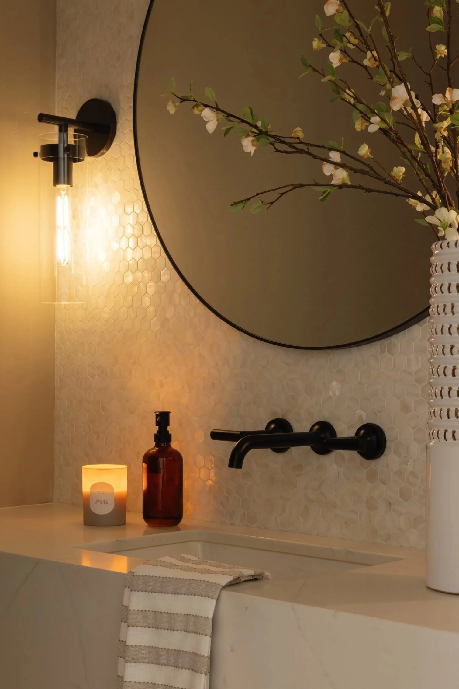 Floating vanity bathroom sink area with a round mirror, black wall-mounted faucet, a lit candle, a brown bottle, and a white towel with gray stripes hanging over the sink edge. There are decorative small hexagonal tiles on the wall.