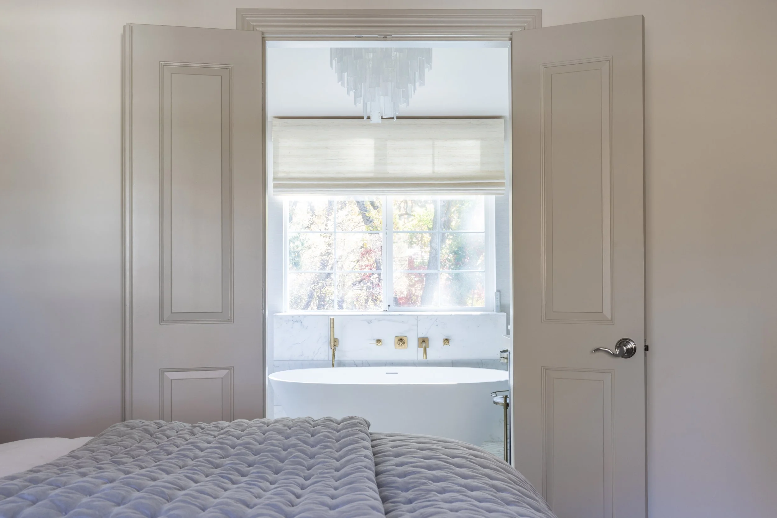 View from a bedroom looking through open double doors into a bathroom with a bathtub, large window with blinds, and chandelier, with part of a bed in the foreground.