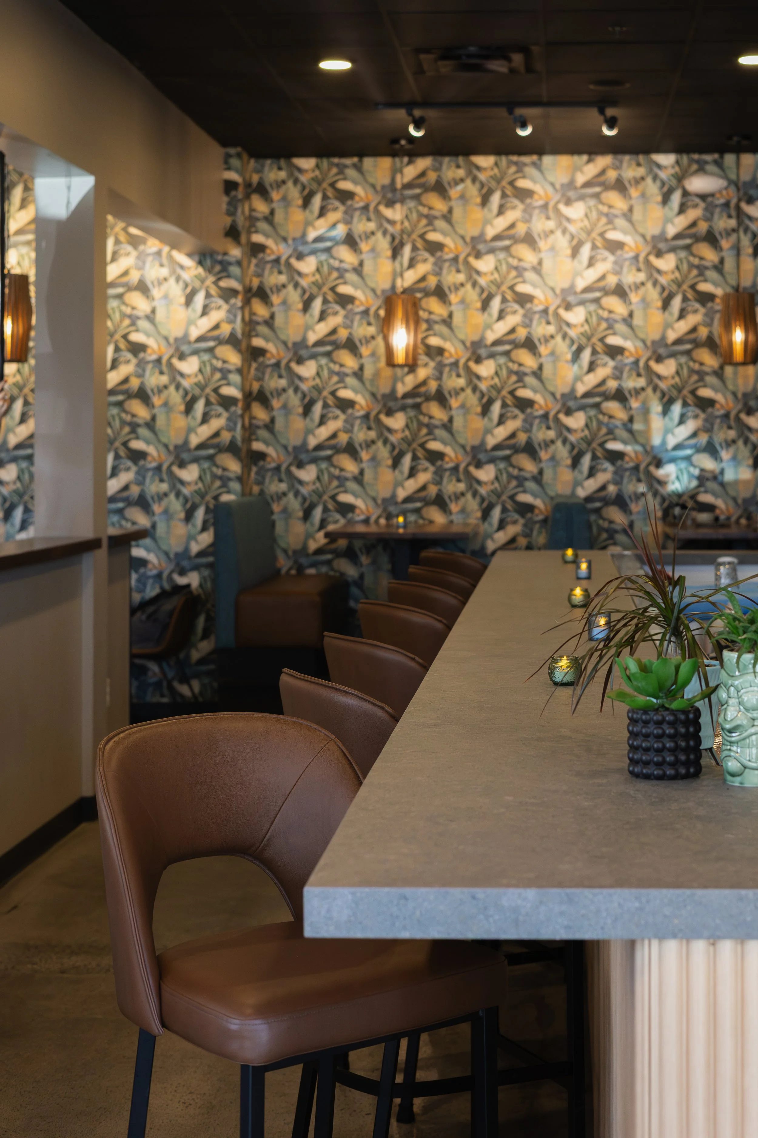 A modern interior with a high counter table and brown chairs, decorated with a patterned wallpaper of floral and leaf motifs, and accented with wall-mounted lamps and small candles.
