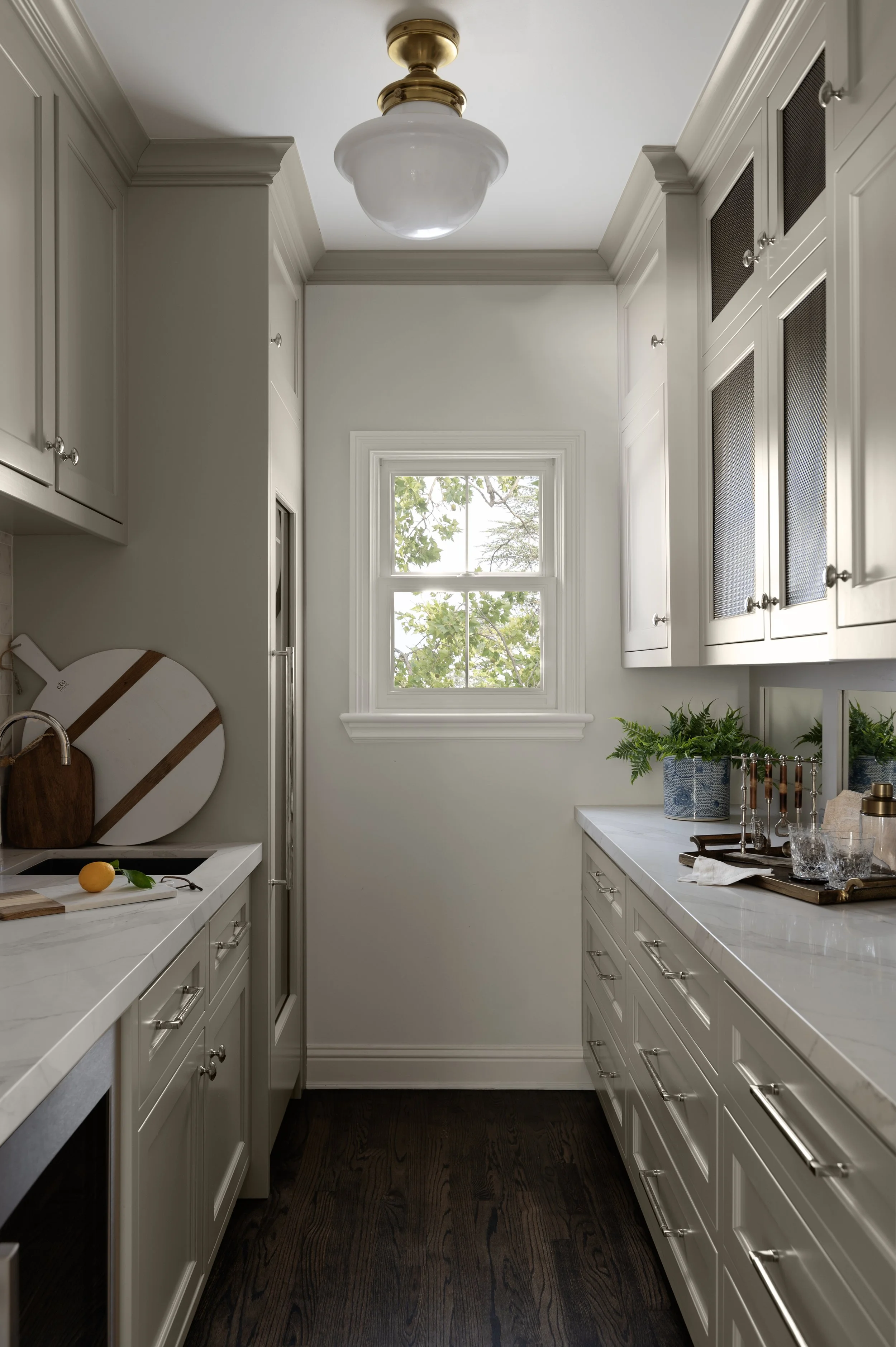A narrow kitchen with white cabinets, dark wood floor, and a window showing green leaves outside. There is a white counter with a cutting board, lemon, and herbs, along with glass jars and a utensil holder.