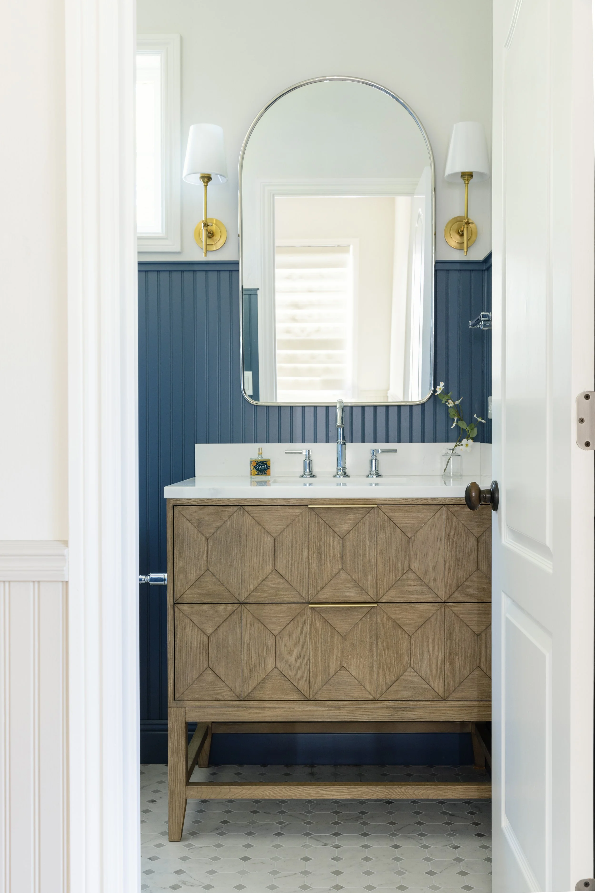 Bathroom vanity with wood cabinet, white countertop, silver faucet, large mirror, blue wall paneling, and two wall-mounted lights, with a small flower in a glass vase.