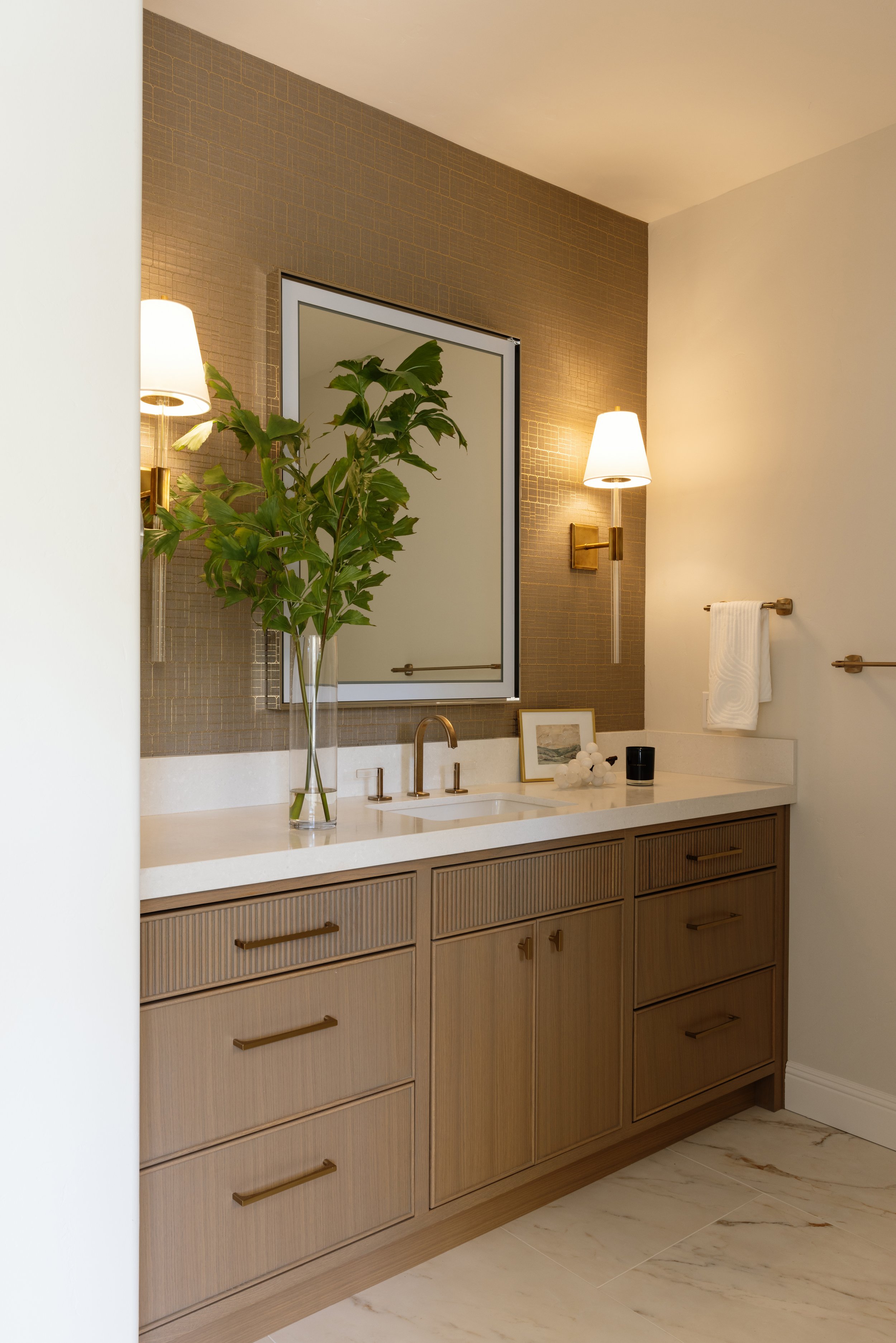 A bathroom vanity with a beige wood finish, gold hardware, a white countertop, and a gold faucet. Behind the sink, there is a large mirror, a vase with green leaves, and framed artwork. On the wall, there are two gold wall sconces with white lampshad