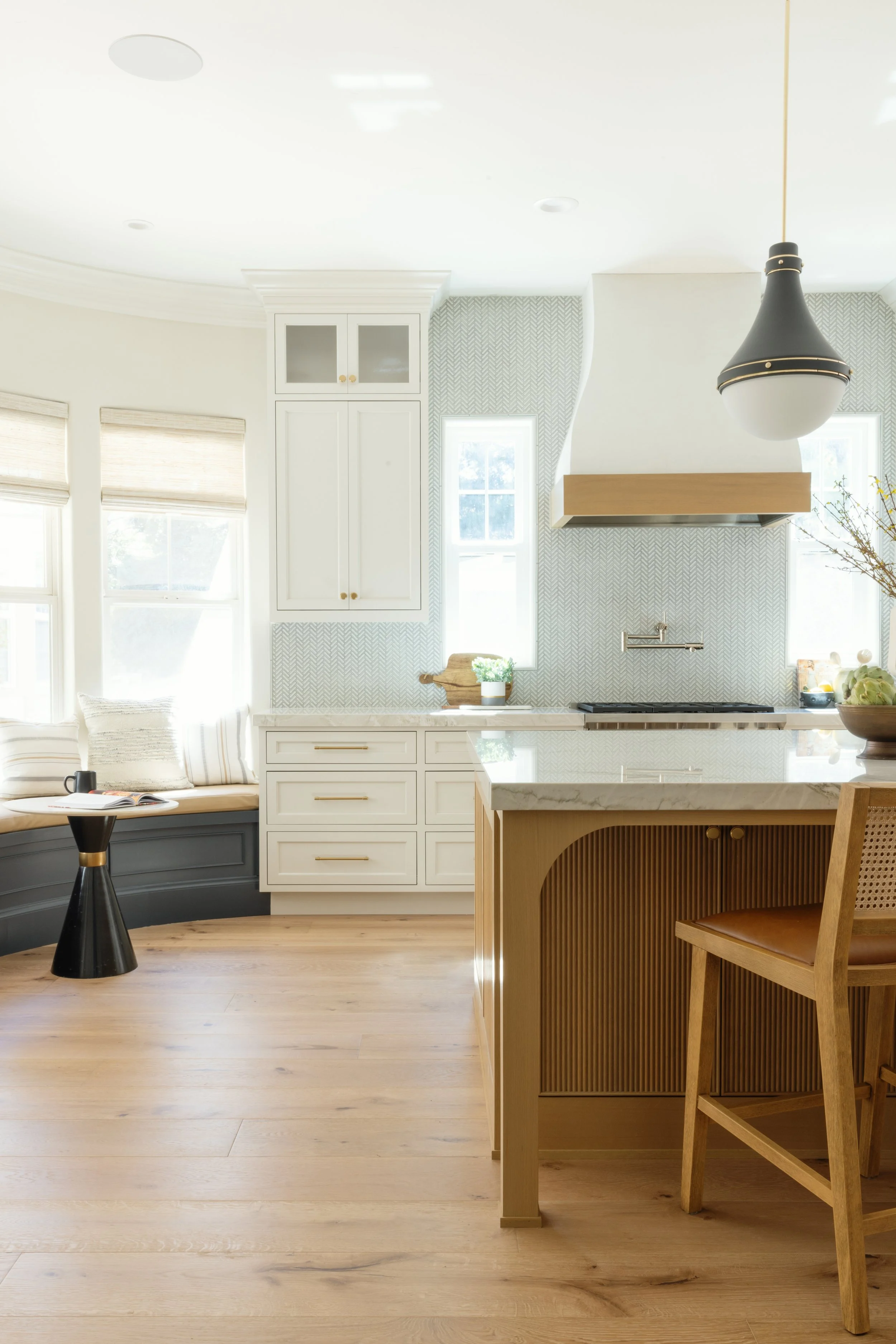 A modern, bright kitchen with light-colored wooden floors, white cabinets, a marble countertop, and a kitchen island with a wooden base. There are two windows with blinds allowing natural light, a black and gold pendant light, and some decorative pla