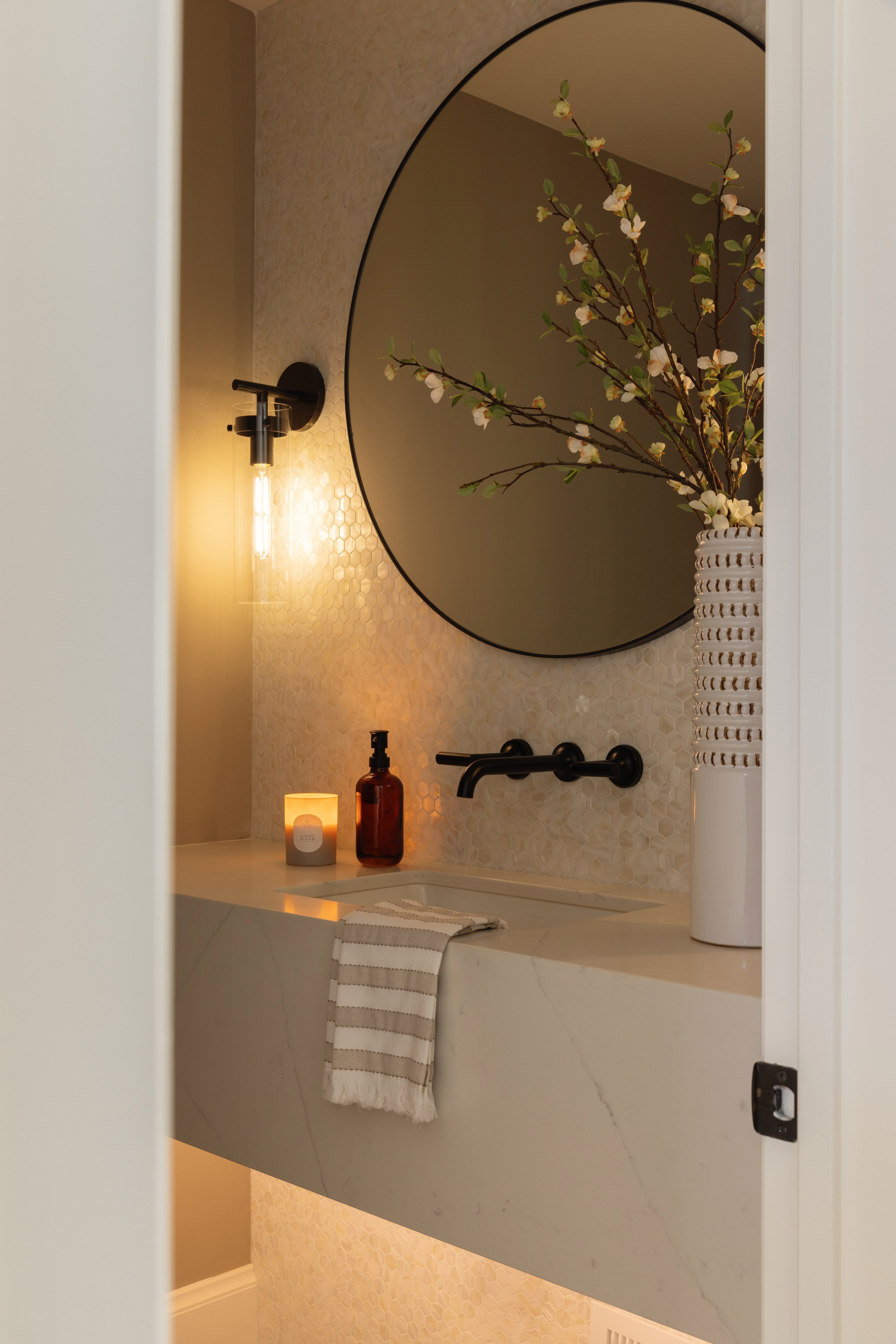 Bathroom vanity area with a round mirror, a black wall sconce with an Edison bulb, a vase with flowers, a candle, a soap dispenser, and a towel hanging over the sink. The wall has textured tile, and the vanity counter is marble.