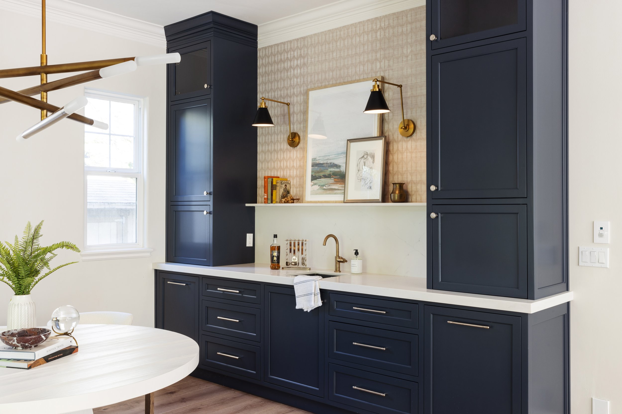 Modern kitchen with navy blue cabinetry, white countertop, brass wall sconces, artwork, a window, and a round white table with books, a decorative bowl, and a glass orb.