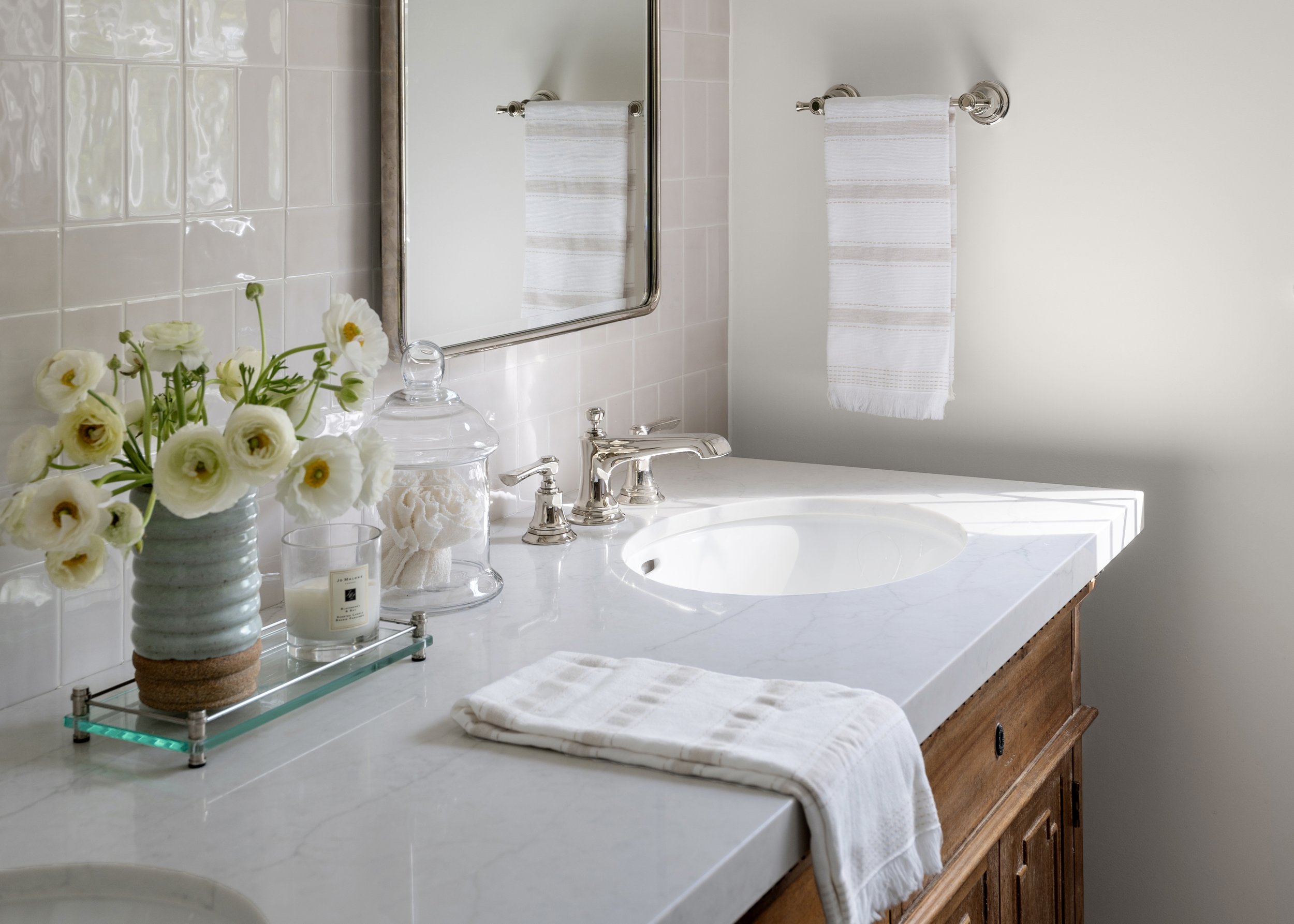 Bathroom vanity with a white marble countertop, a rectangular mirror, a vase of white flowers, candles, and neatly folded towels, with a white wall and a towel hanging on a chrome bar.