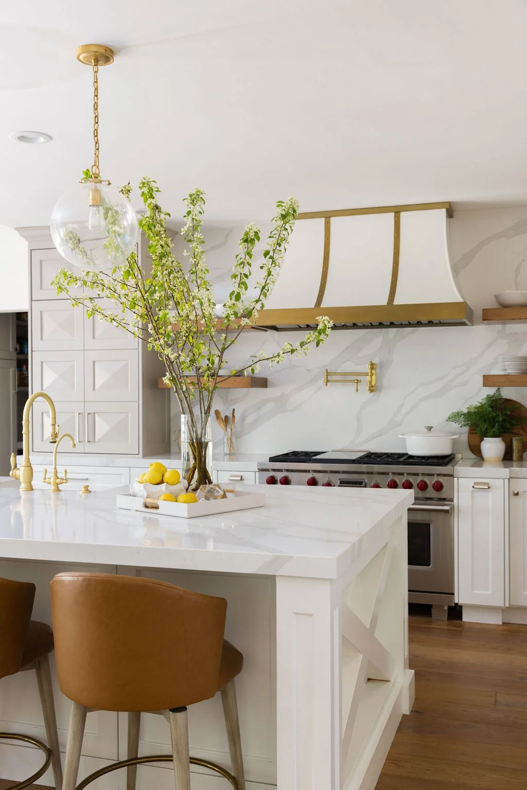 A modern kitchen with white cabinets, a white marble countertop, a large glass vase with tall green branches and white blossoms, a tray of lemons, and gold accents including a faucet, light fixture, and range hood. There are brown chairs at the count