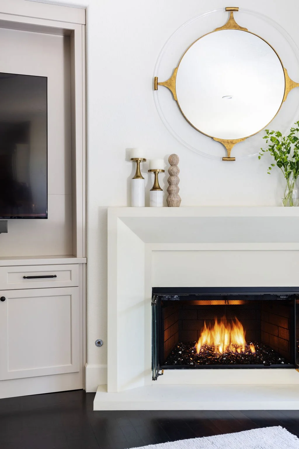 Living room with white fireplace, burning fire, modern decor, gold-framed circular mirror, white cabinets, decorative candles, and green plant.