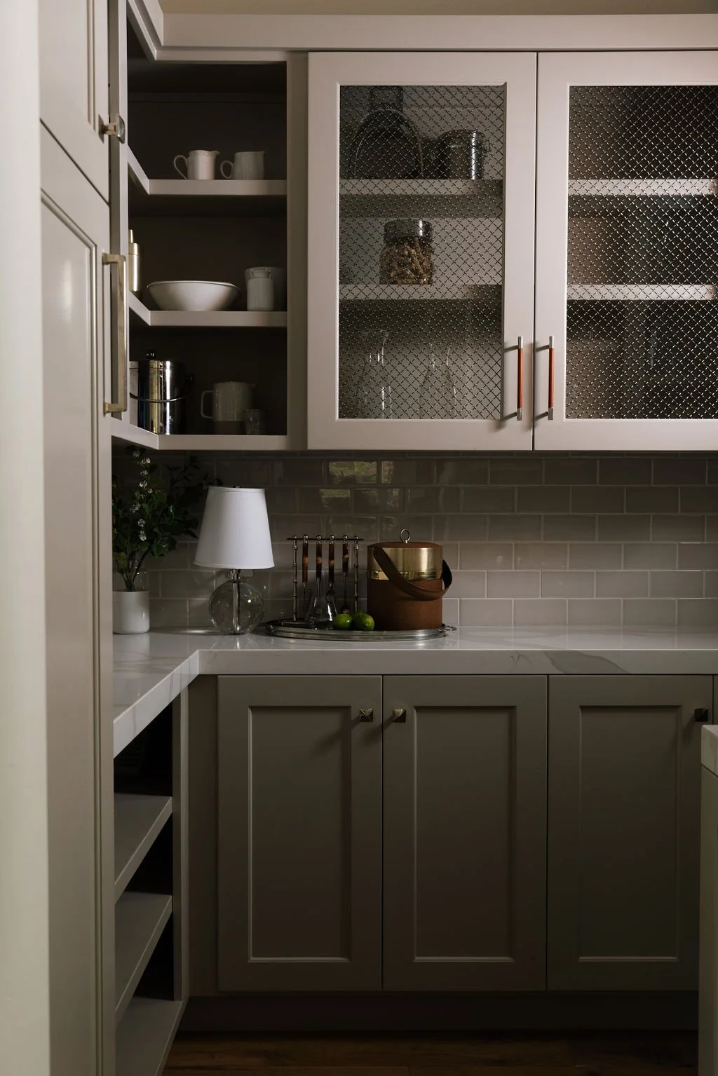 Kitchen cabinet with open and closed shelves, containing dishes, glasses, and jars; counter has a lamp, a plant, and a tray with limes and a container.