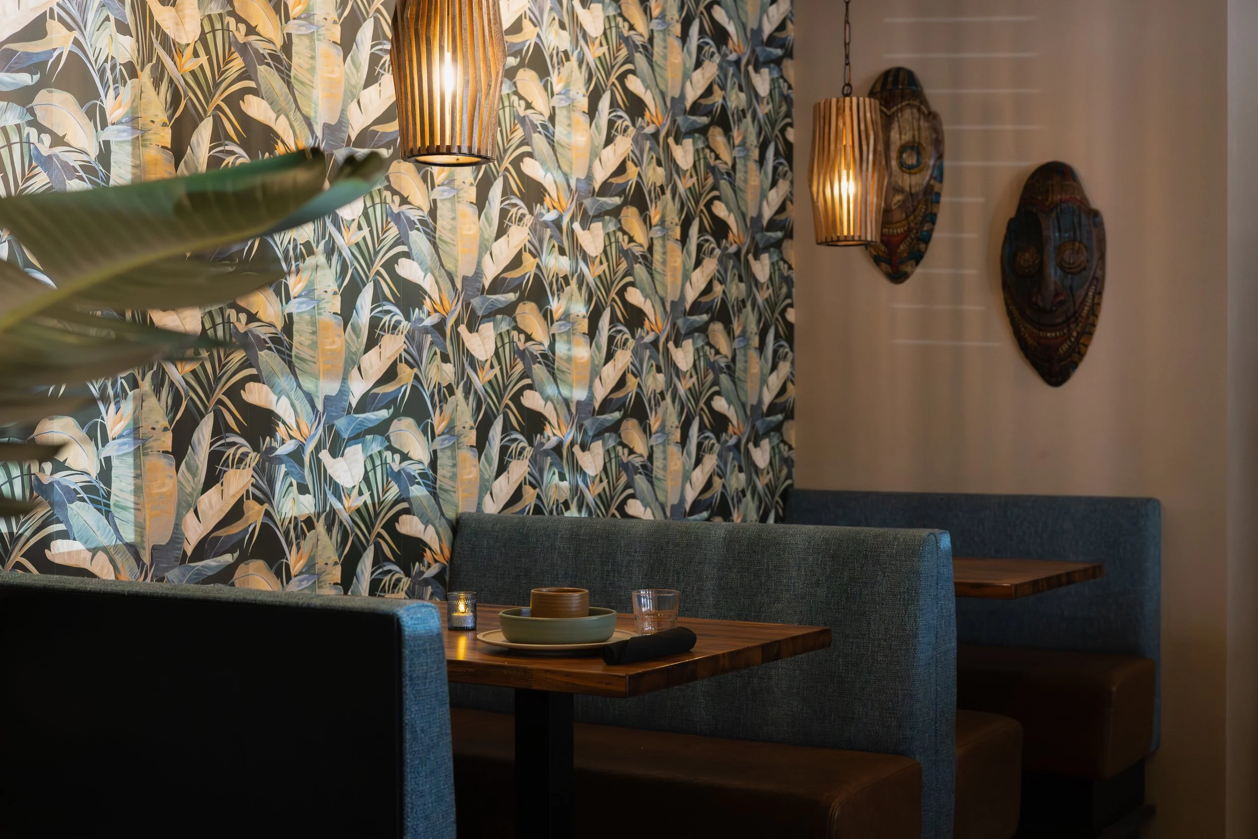 Interior of a restaurant or cafe with a tropical leaf patterned wallpaper, three hanging pendant lights with wooden slats, a wall decoration with African masks, a dark upholstered booth, tables with dishes, glasses, and lit candles.