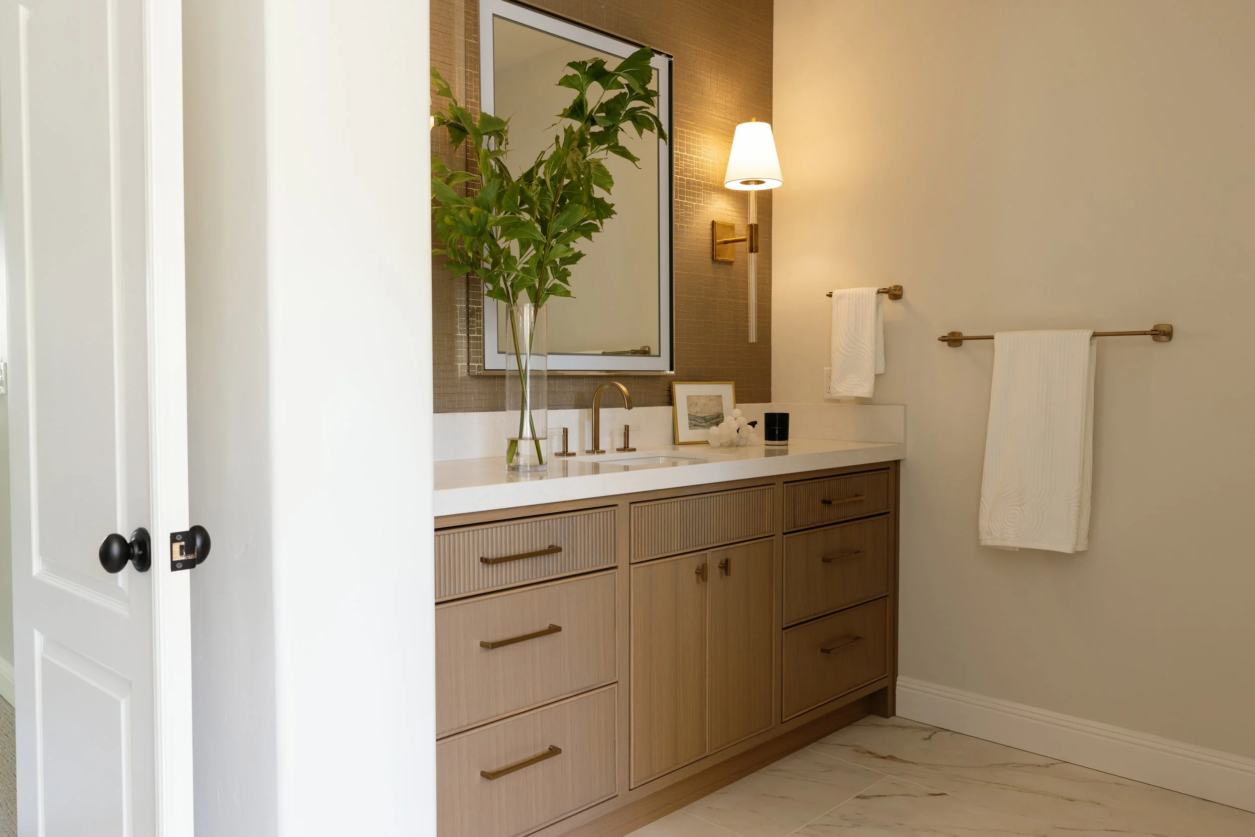 Modern bathroom vanity with wood cabinetry, a white countertop, and a large mirror; decorated with a tall green plant, a framed picture, and a small black object, with a wall-mounted lamp and towel holder with towels.