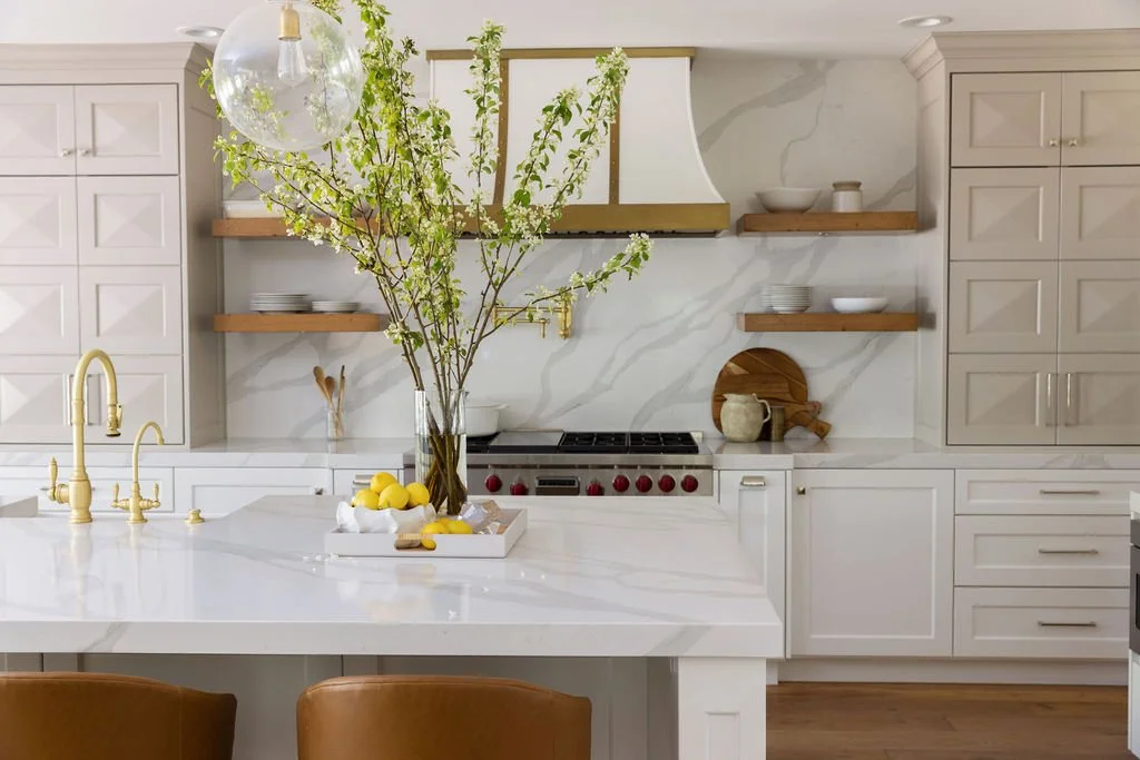 Modern kitchen with white cabinets, a marble backsplash, open wooden shelves, and a large island with a white marble countertop. A tall glass vase with green branches and lemons sits on the island, along with a bowl of lemons. Gold fixtures are on th