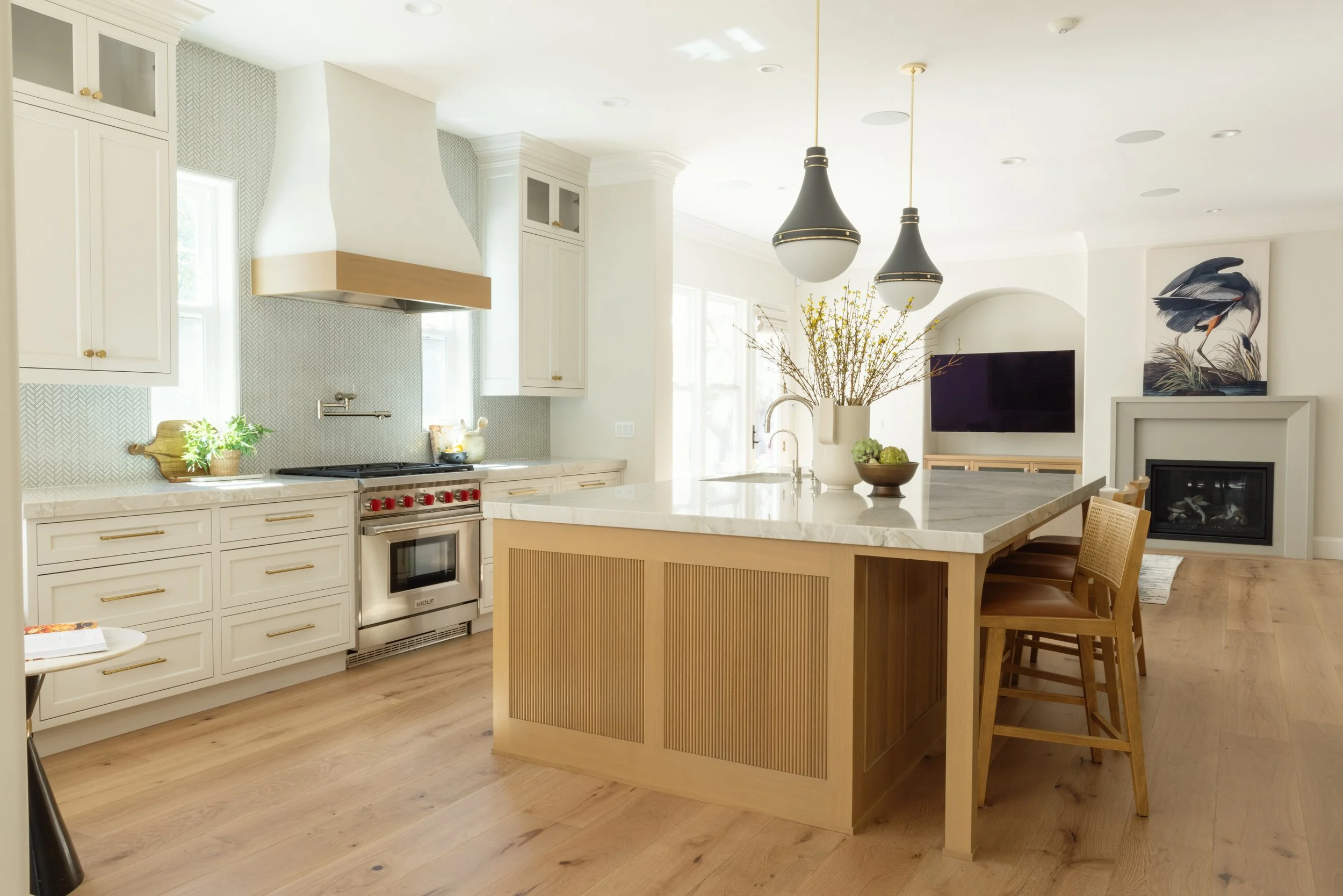 Modern tuscan kitchen remodel with white cabinets, marble countertops, and a large island with reeded wooden paneling. There are hanging pendant lights, arched island legs. Floating hood with Ann Sacks Savoy herringbone tile