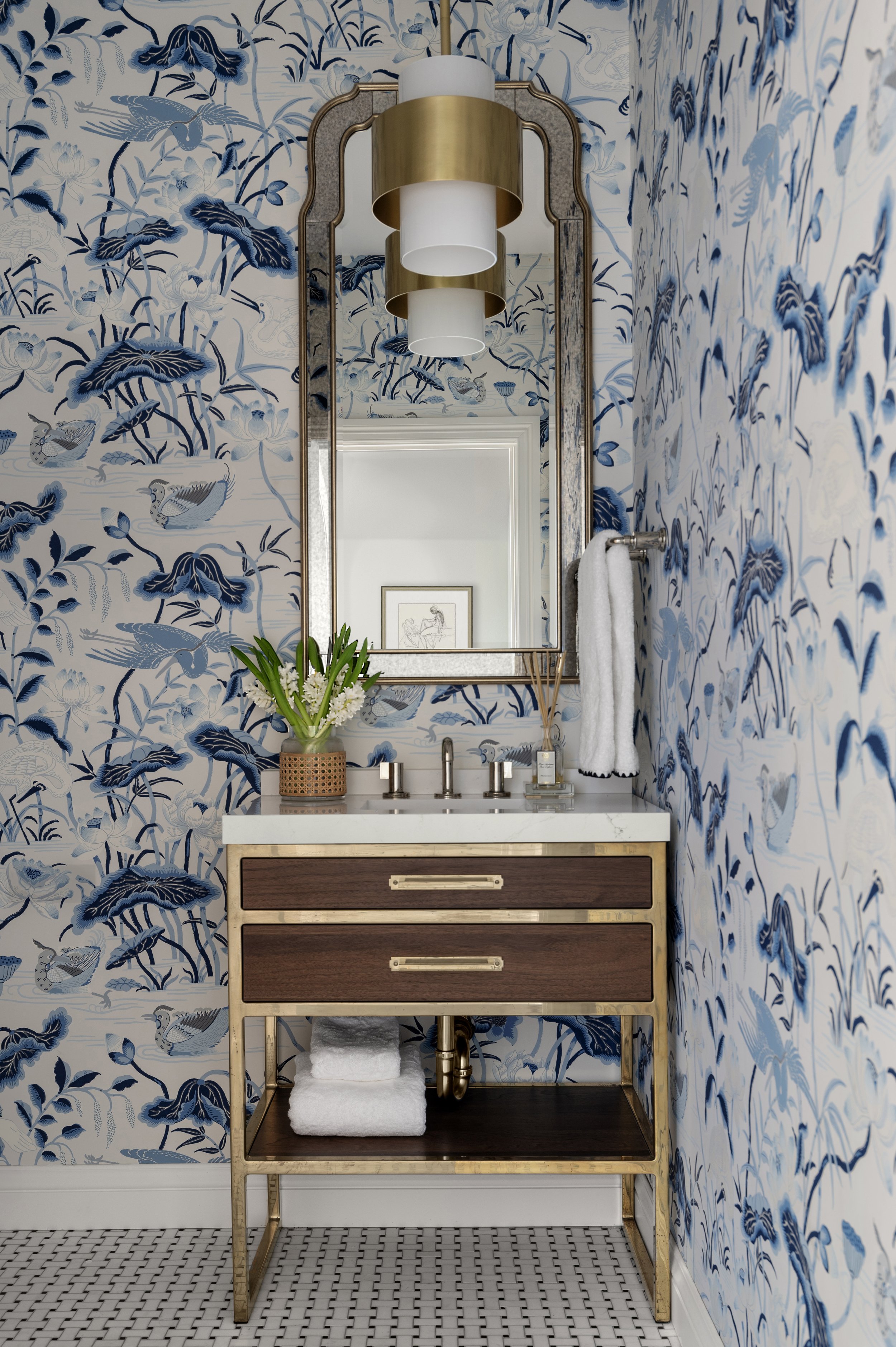 Bathroom vanity with a wooden cabinet and gold frame, topped with a white marble countertop, set against walls with blue and white floral wallpaper featuring birds, with a wall-mounted mirror, a vase with white flowers, a soap dispenser, and a stacke