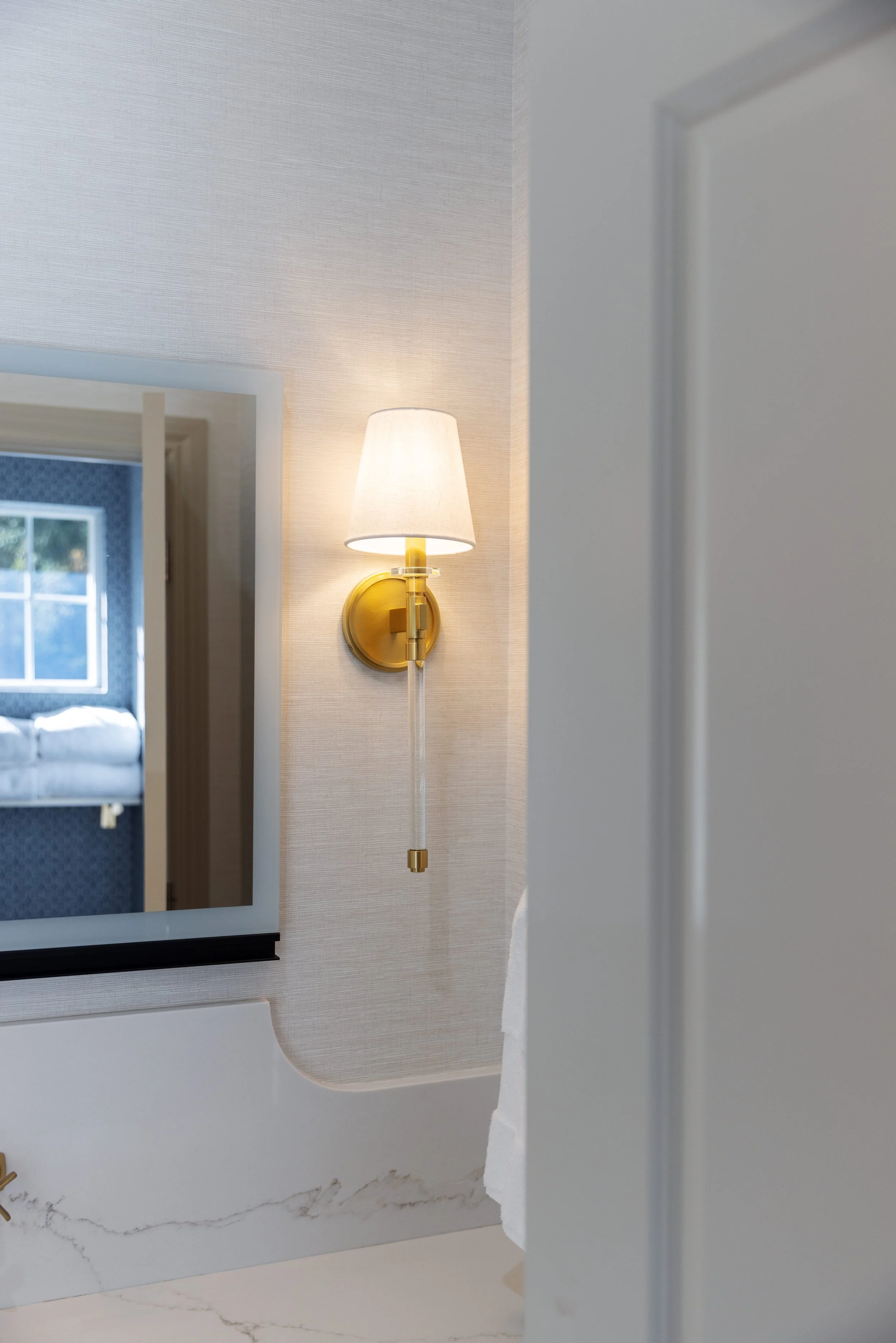 Interior view showing a wall-mounted gold and white lamp next to a mirror, with a doorway leading to a room with blue wallpaper and a bed, and a marble-like surface at the bottom.