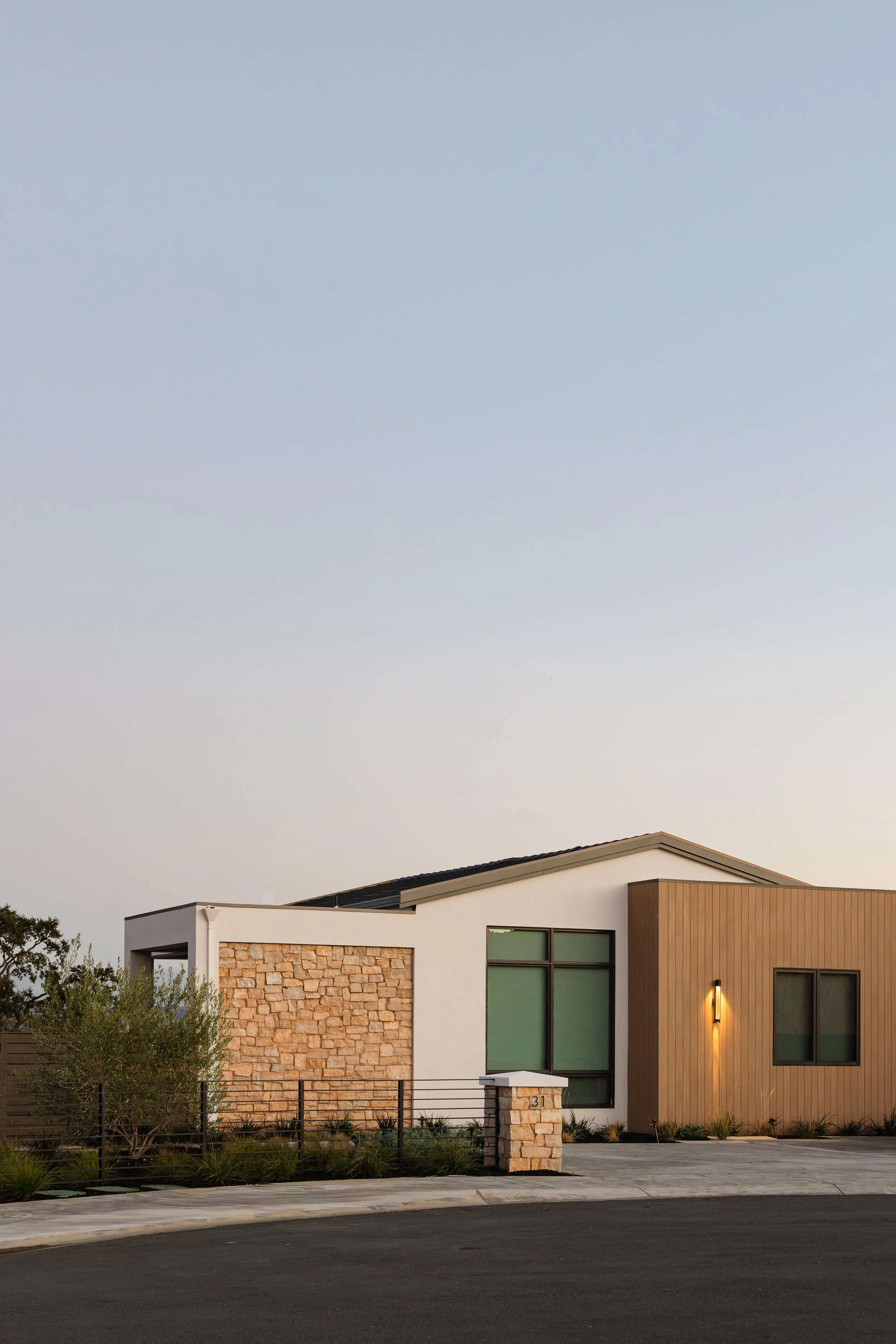 Modern house with a mixture of materials including white stucco, stone, and wood paneling, large windows, and a front yard with plants and a stone mailbox, taken at dusk.