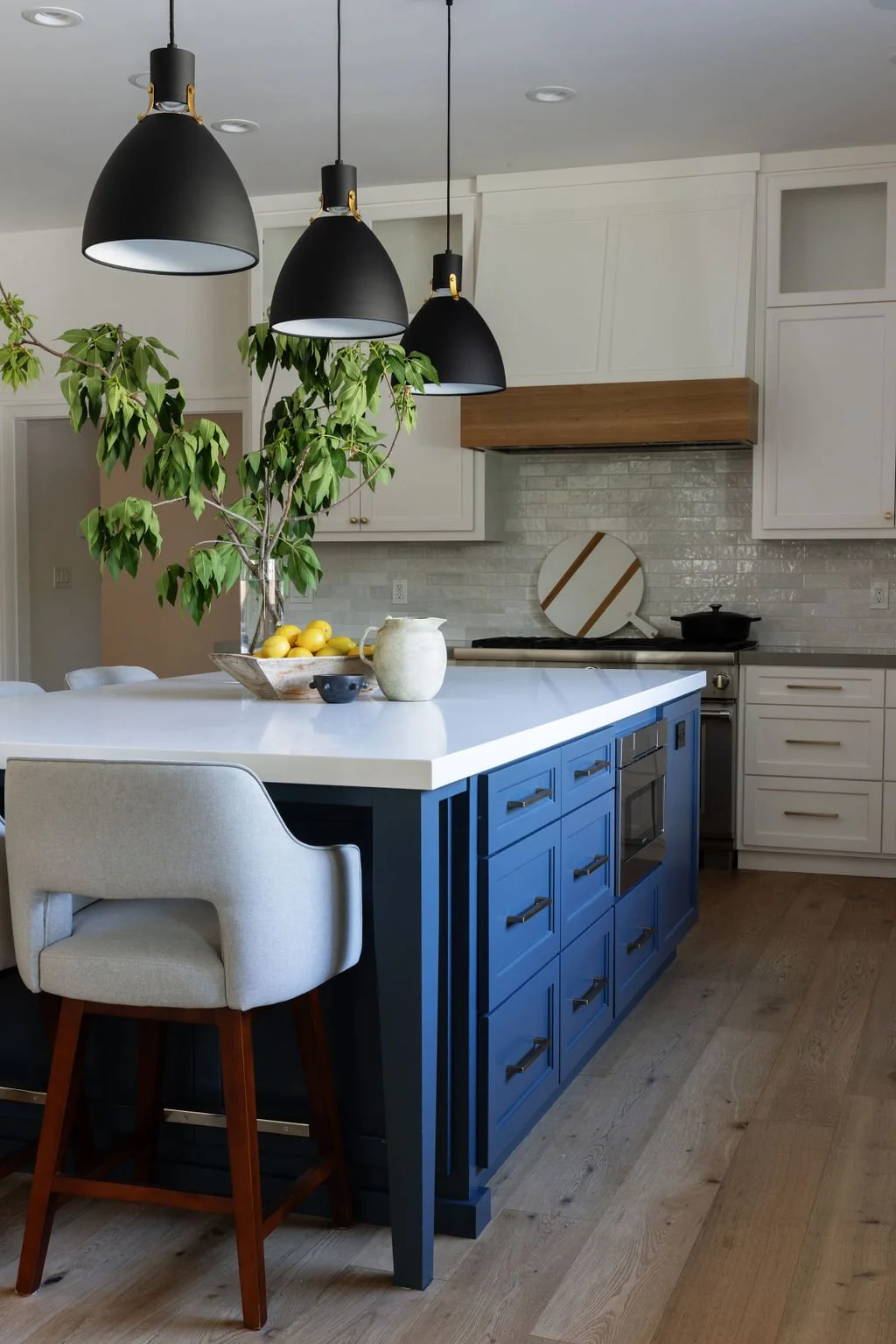 Modern kitchen with white cabinetry, a blue kitchen island, black pendant lights, a large potted plant, and decorative items on the island including a bowl of lemons, a white pitcher, and a small black bowl.