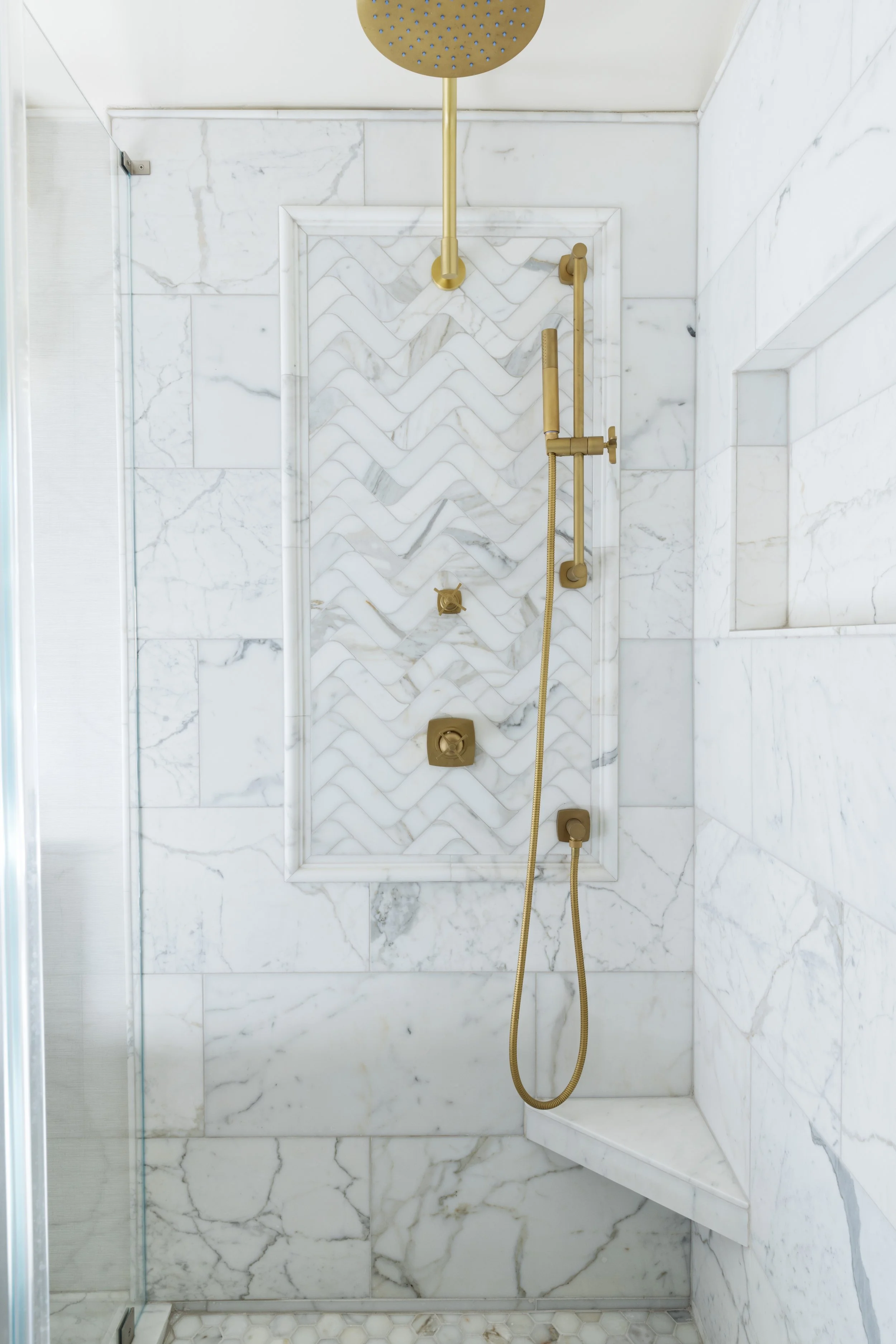 A luxurious walk-in shower with white marble walls and a decorative tile pattern on the shower wall. It features a gold showerhead, a hand-held shower, and gold fixtures, with a glass panel on the left side.