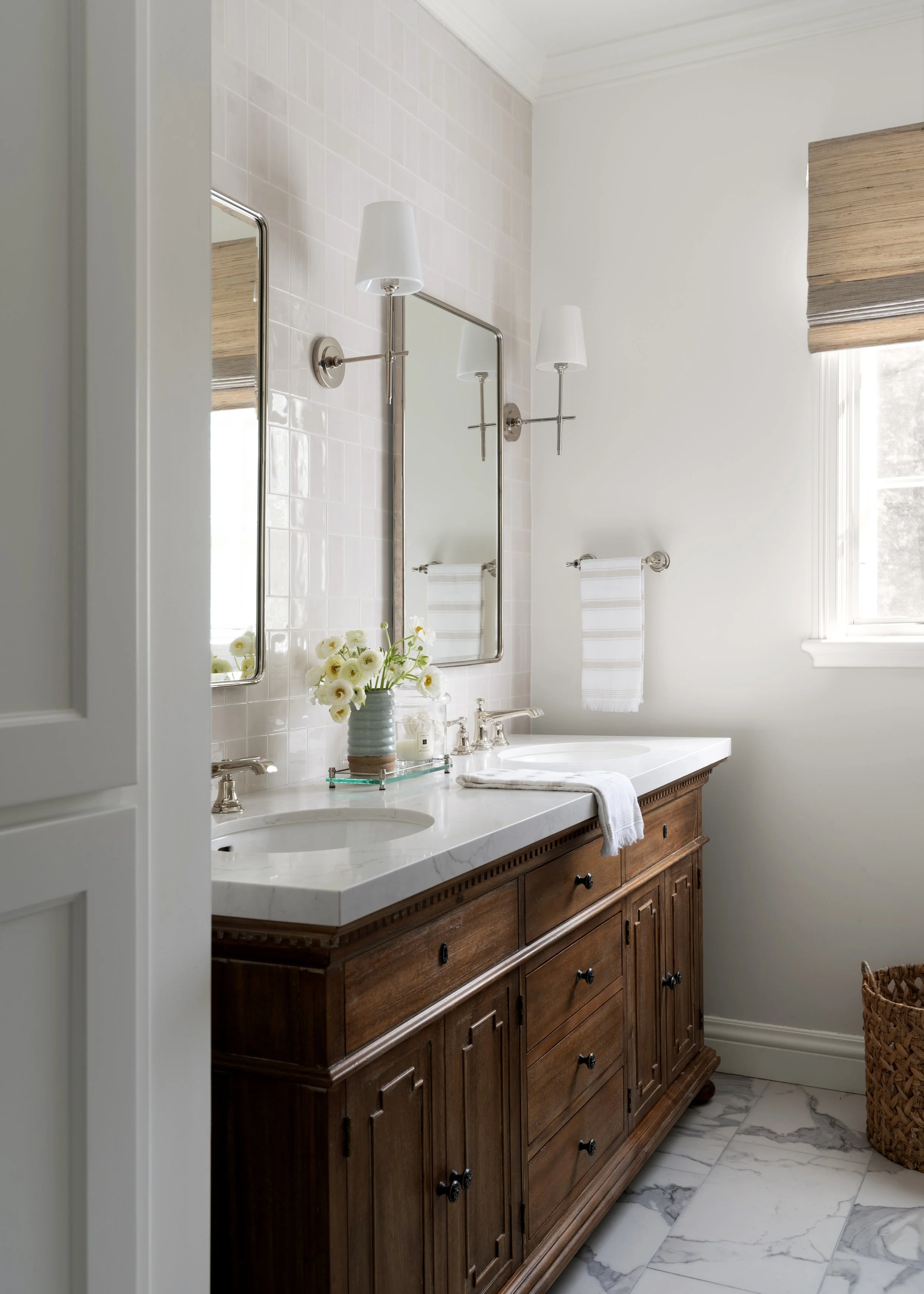 A bathroom vanity with a white marble countertop, a double sink, and a wooden cabinet base, with a vase of white flowers, a mirror, and wall-mounted lamps reflected in the mirror.
