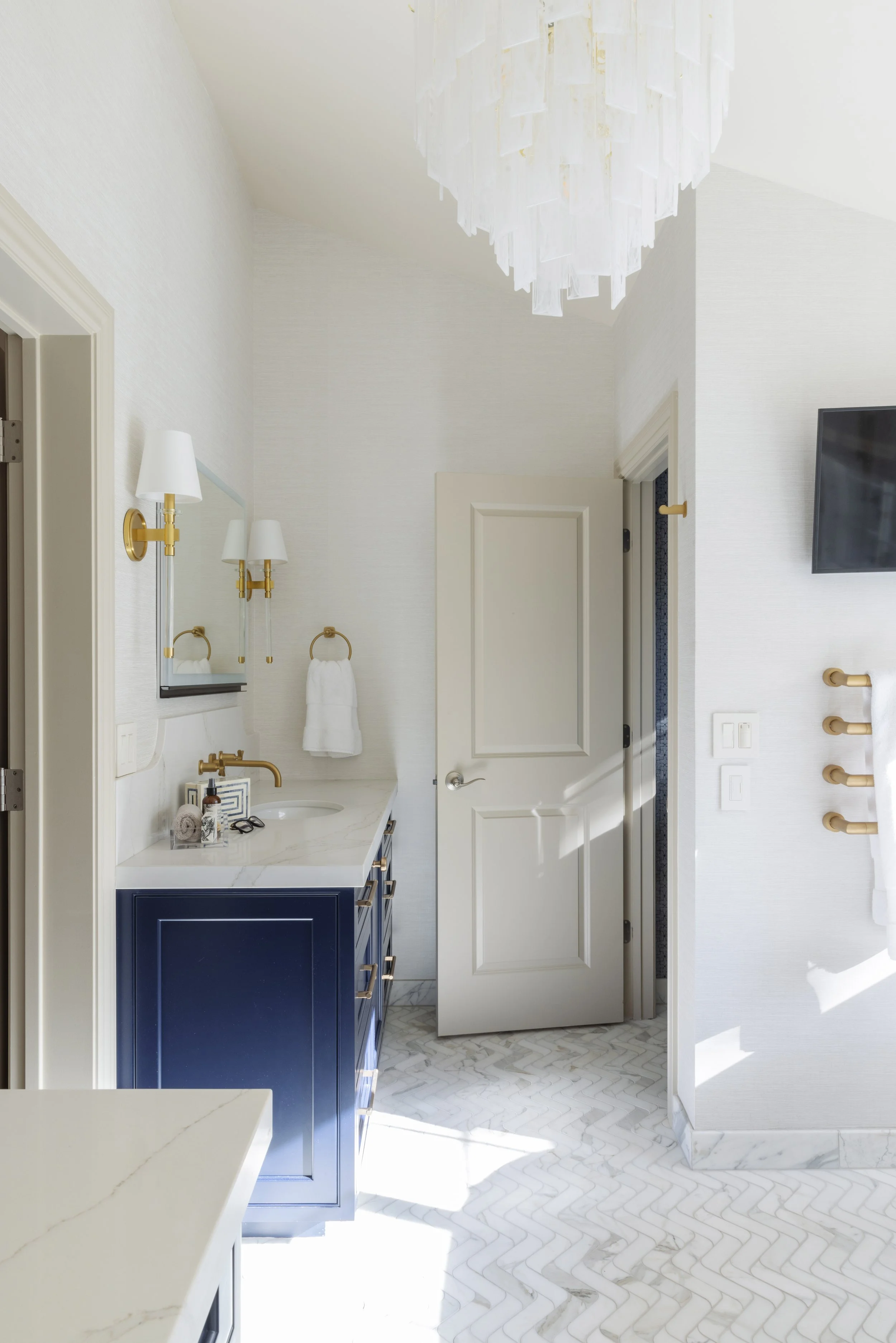 A bright, modern bathroom with white walls, a navy blue vanity with gold handles, white marble countertop, gold fixtures, a white towel hanging on a ring, a mirror with gold wall sconces, and a white door. Light streams in, illuminating marble herrin