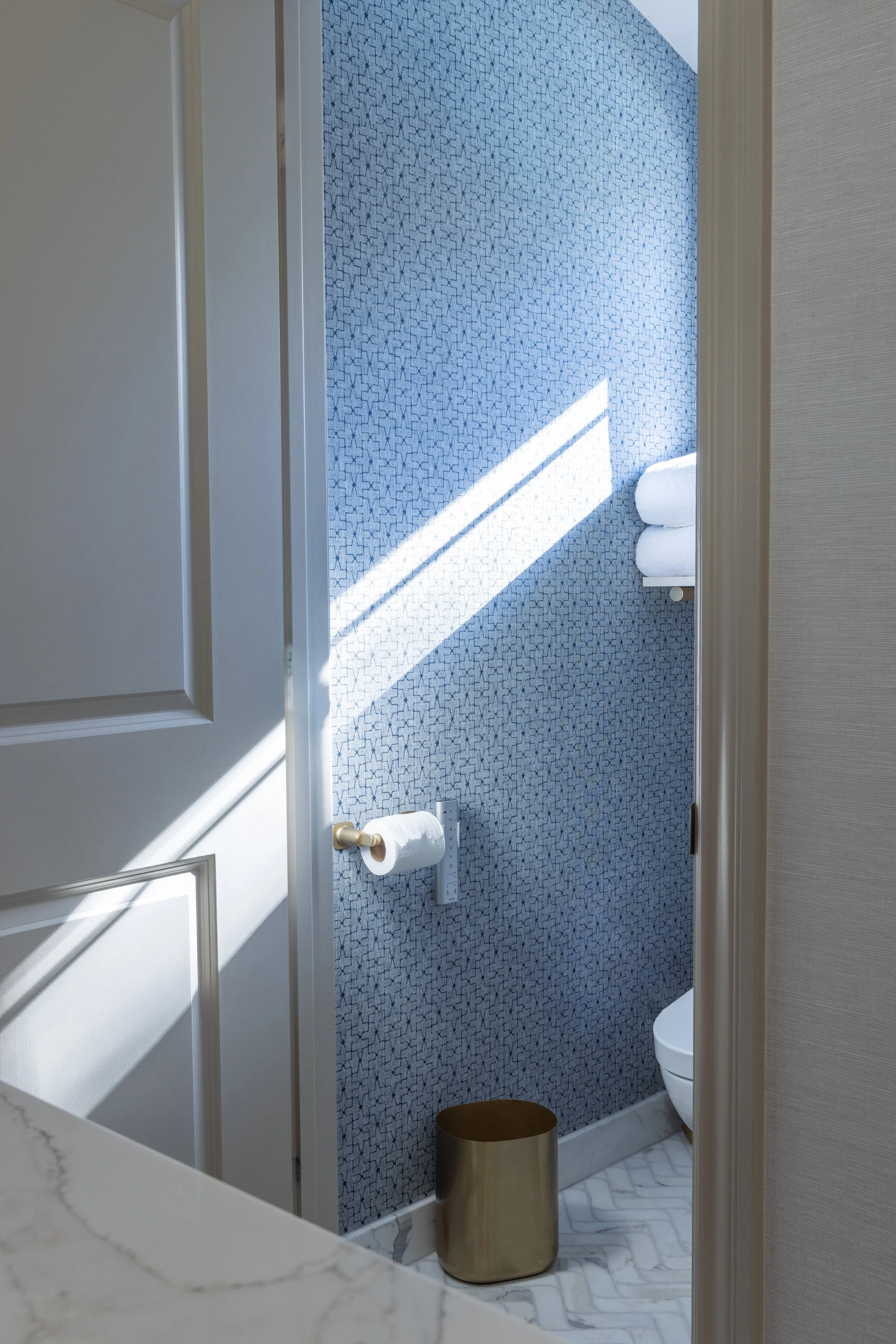 A bathroom with a patterned blue wall, white towels stacked on a shelf, a white toilet, and a gold-colored cylindrical trash can on a white marble floor. Sunlight creates a diagonal shadow across the blue wall.